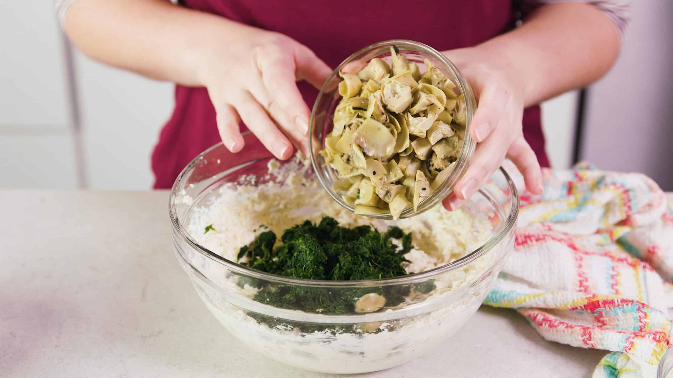 Angled view of a large clear glass mixing bowl filled with cheesy dip mixture with frozen spinach, thawed and drained with artichoke hearts being poured from above from a clear glass mixing bowl.