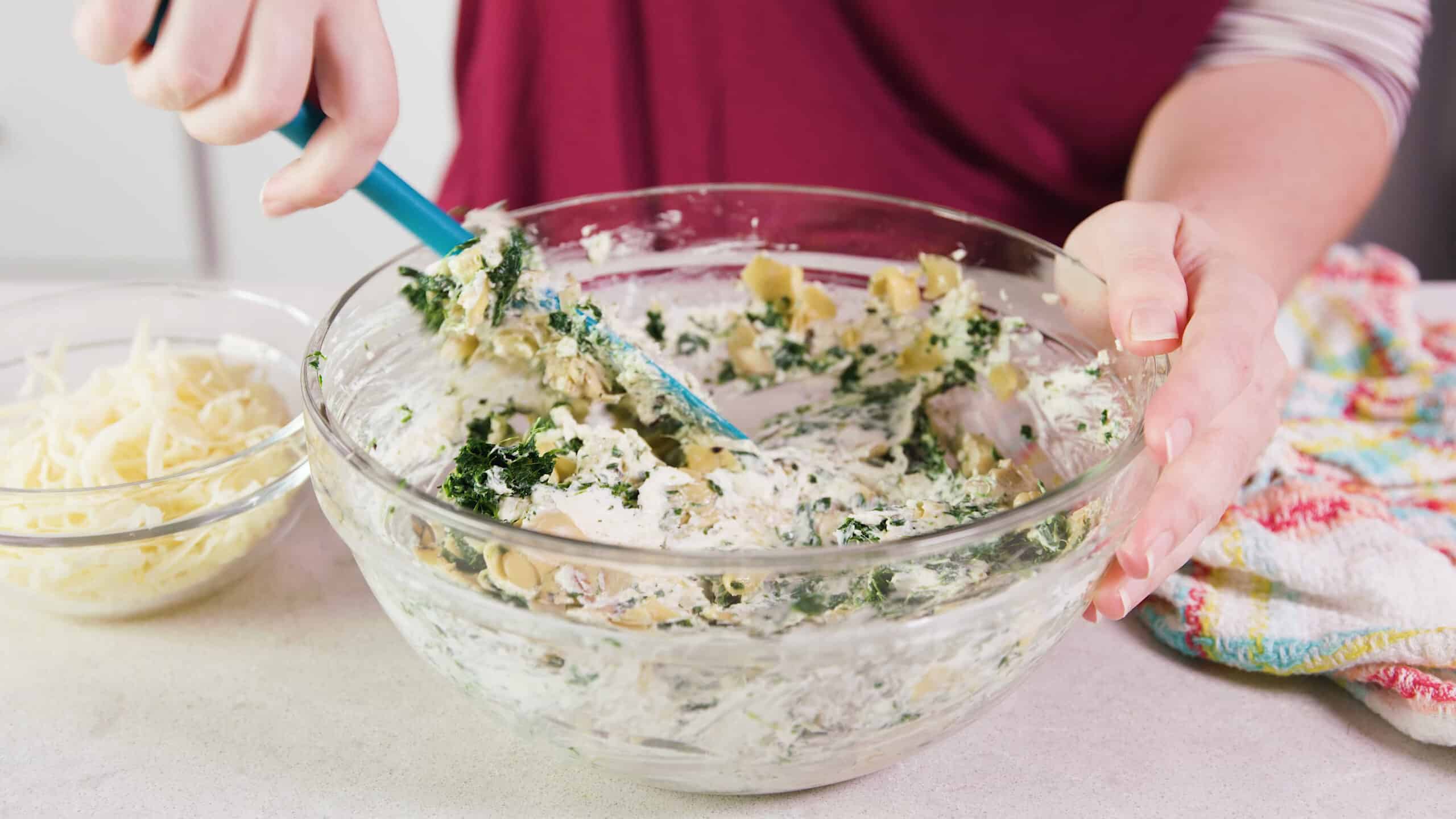 Angled view of a large clear glass mixing bowl filled with cheesy dip mixture, combined with thawed and drained frozen spinach, and artichoke hearts being stirred together with a plastic spatula.