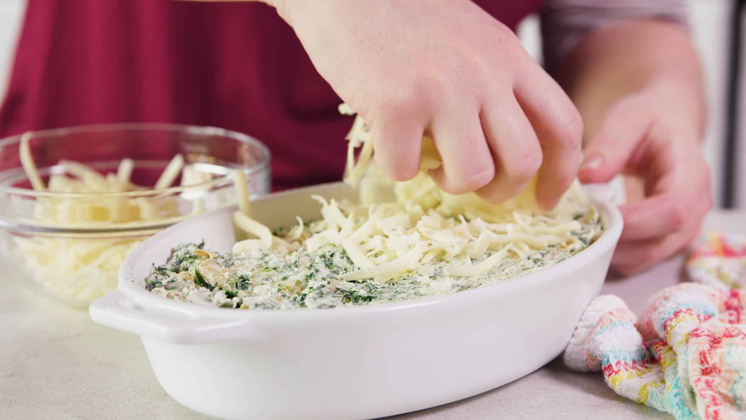 Angled view of white casserole dish filled with cheesy spinach and artichoke dip being topped with shredded mozzarella cheese.