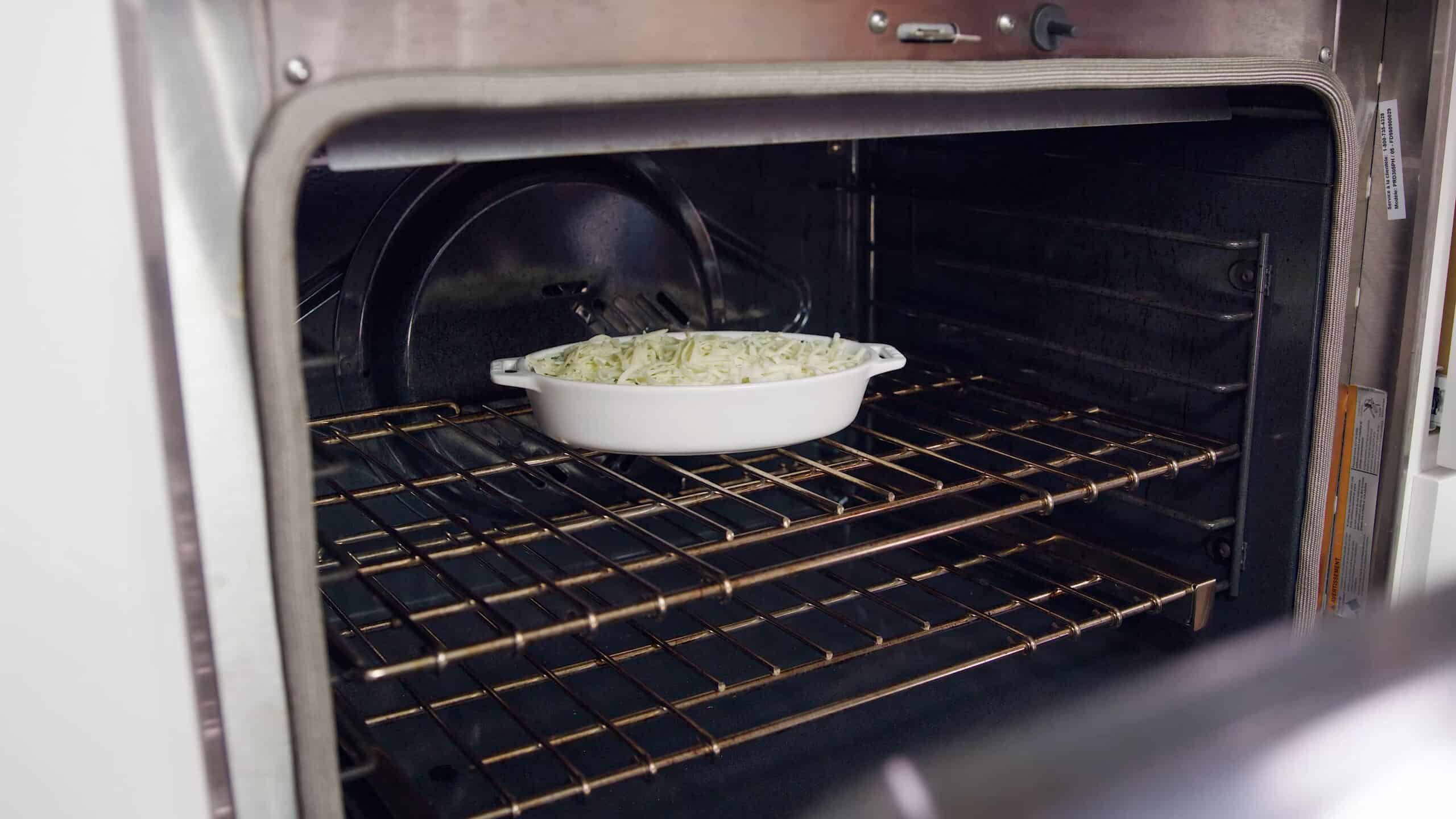 Angled view of an open oven with a white glass casserole dish filled with cheesy spinach artichoke dip placed on a metal rack in the middle of the oven.