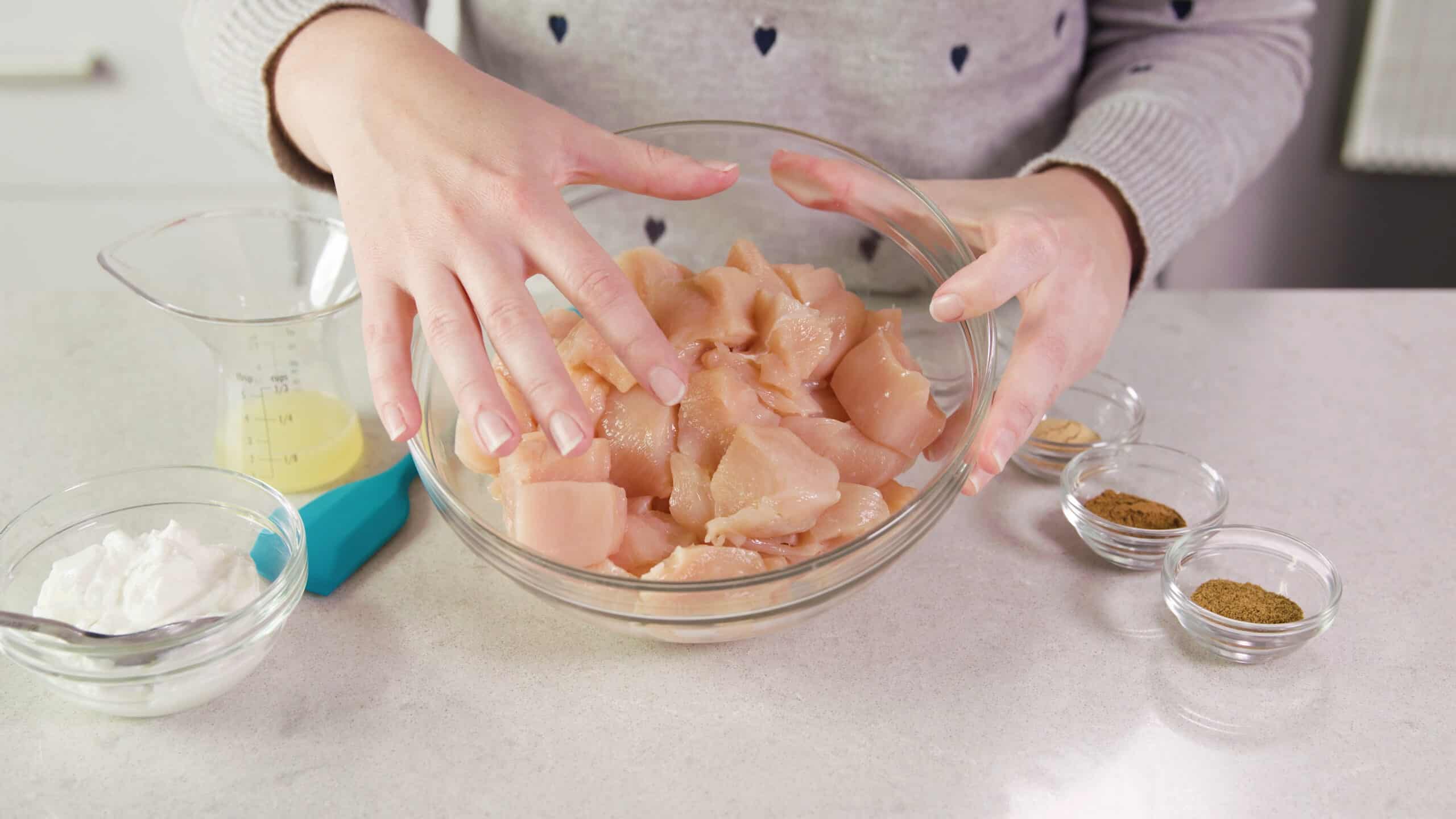 Angled view of clear glass mixing bowl filled with diced raw chicken and dry spices to the right and wet ingredients to the left all on a marble countertop.