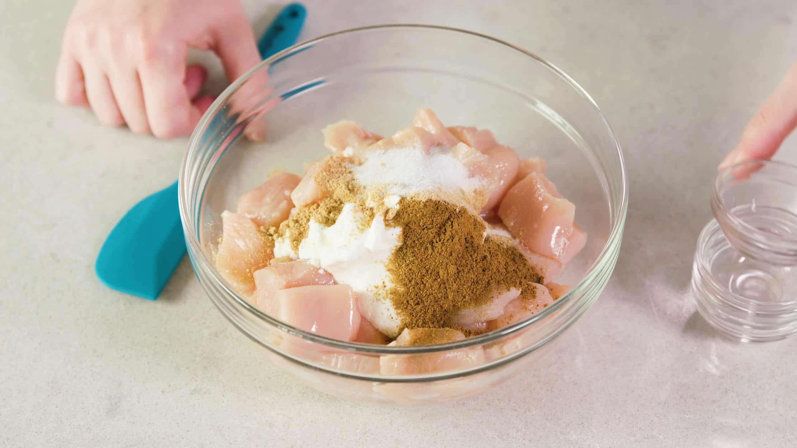 Angled view of clear glass mixing bowl filled with all ingredients and spices for the chicken and marinade ready to be combined with a plastic spatula.