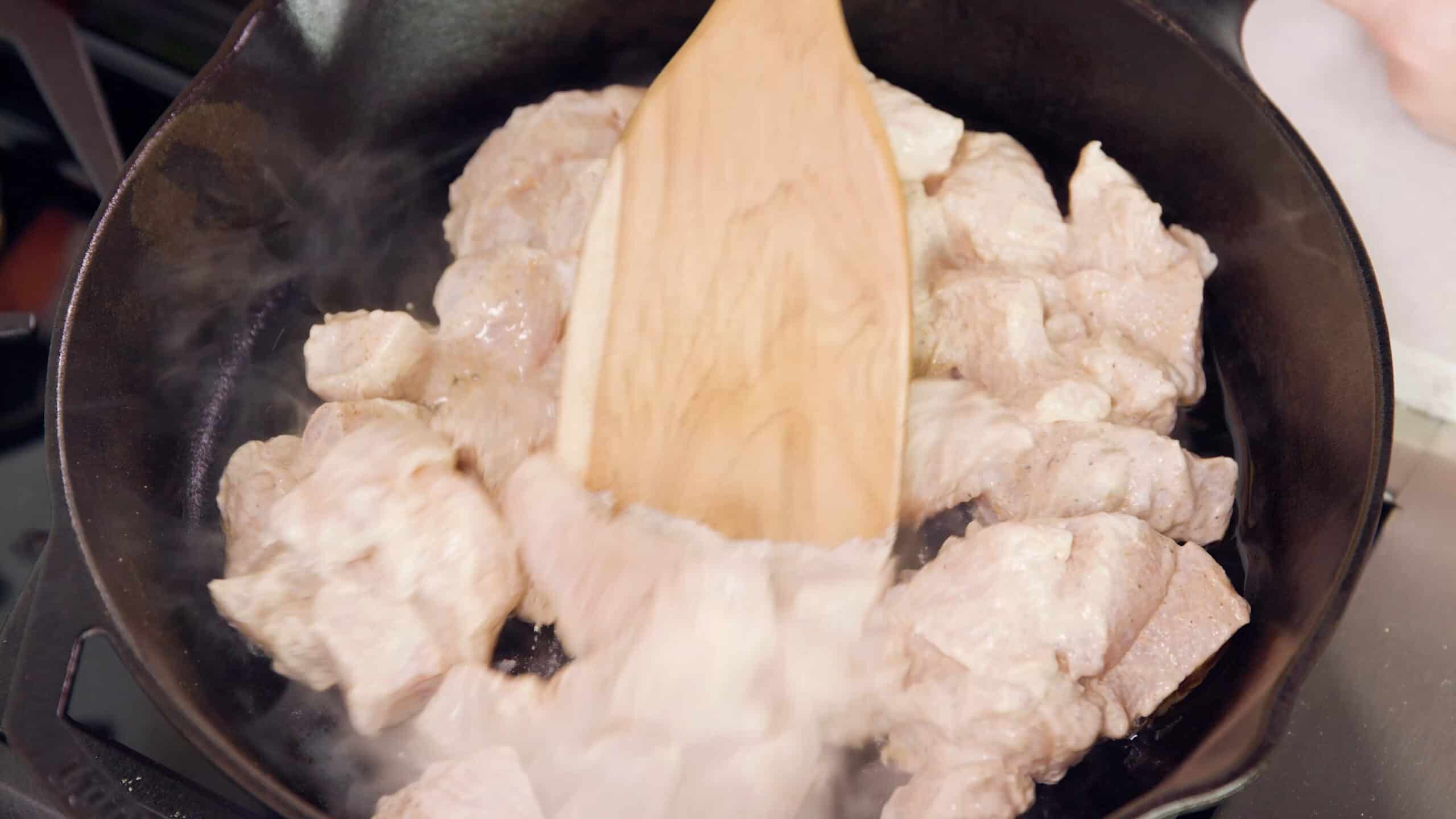 Overhead view of a cast iron skillet filled with sizzling bites of chicken with a wood spatula stirring.