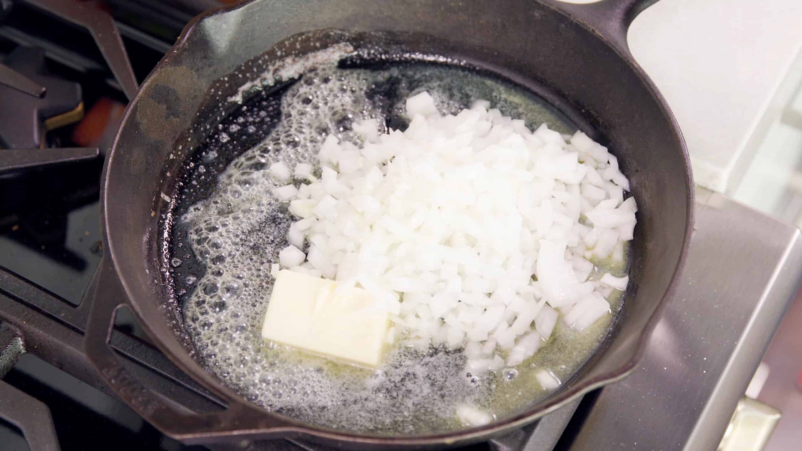 Angled view of a cast iron skillet with white onions and melting butter sizzling on the stovetop.