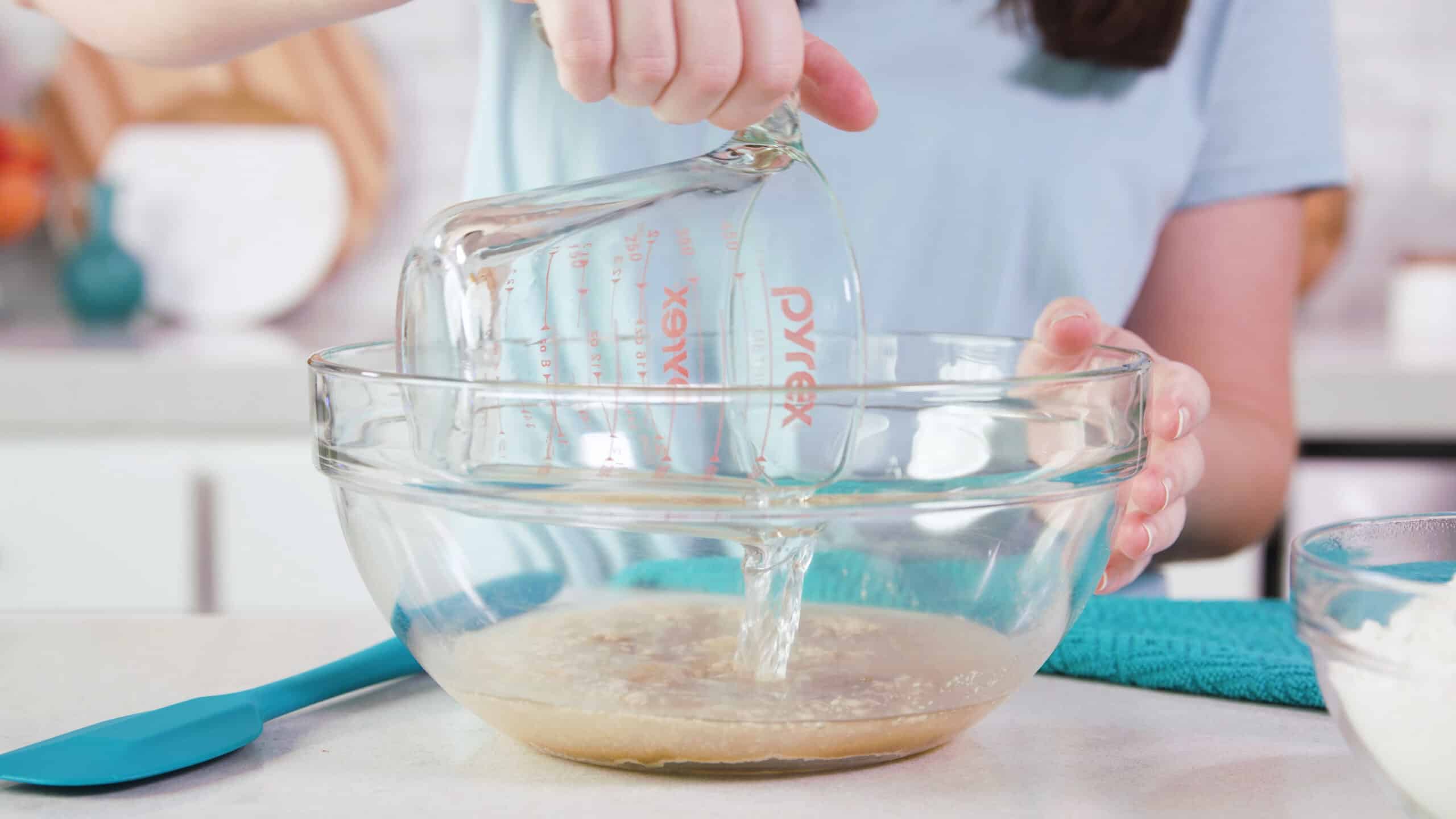 Side view of clear glass mixing bowl filled with dry ingredients and water pouring in from a clear glass measuring cup.