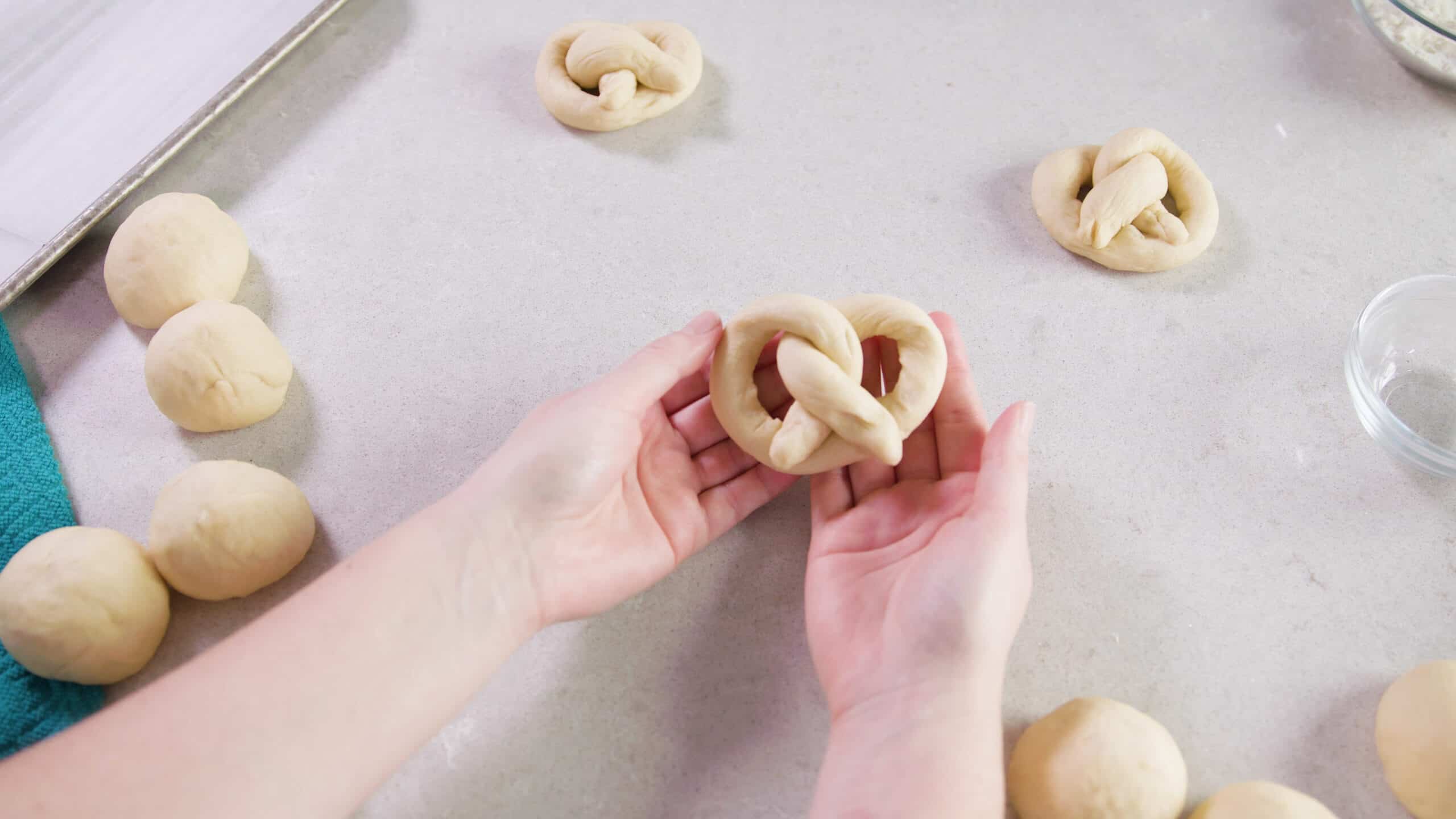 Bird's eye view of clean marble countertop and two hands holding raw dough shaped into a pretzel with other dough balls waiting to be shaped.