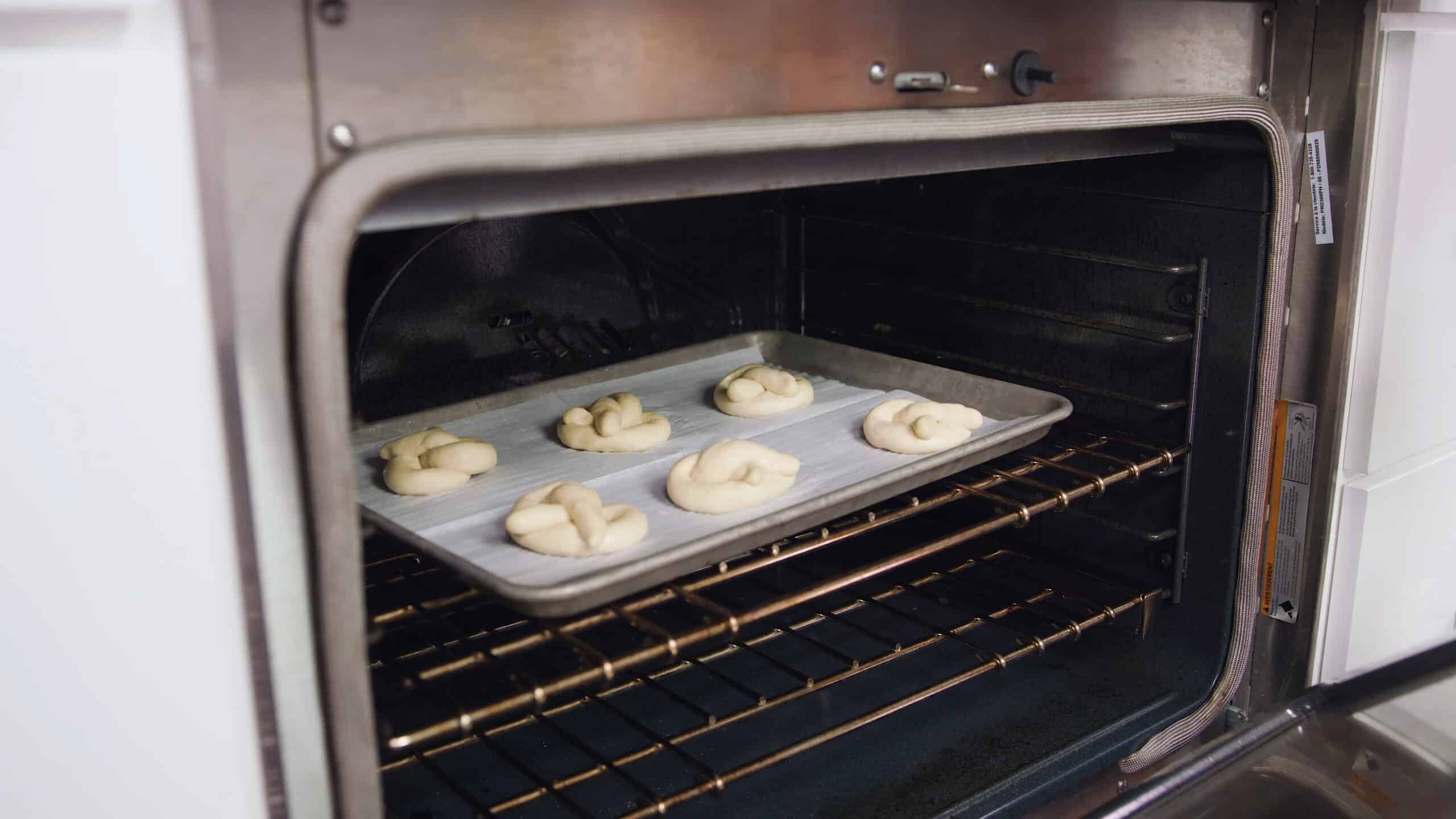 Angled view of open oven with metal baking sheet lined with parchment paper and six hot water dredged shaped pretzels place on a metal rack in the middle of the oven.