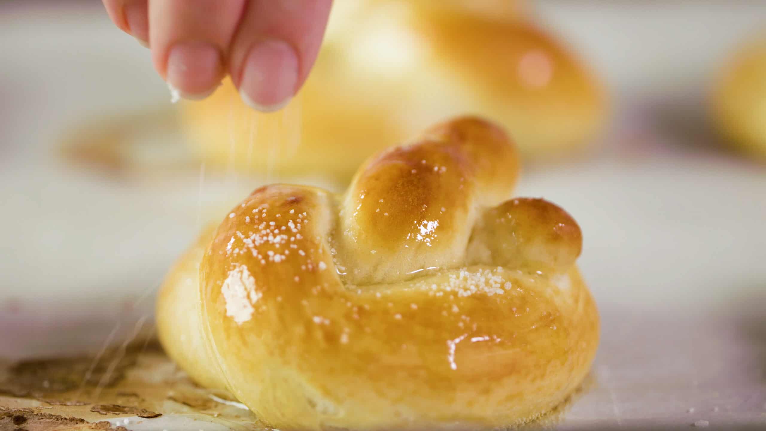 Close-up view of golden brown baked soft pretzel brushed with melted butter and a hand pinching sea salt on top.