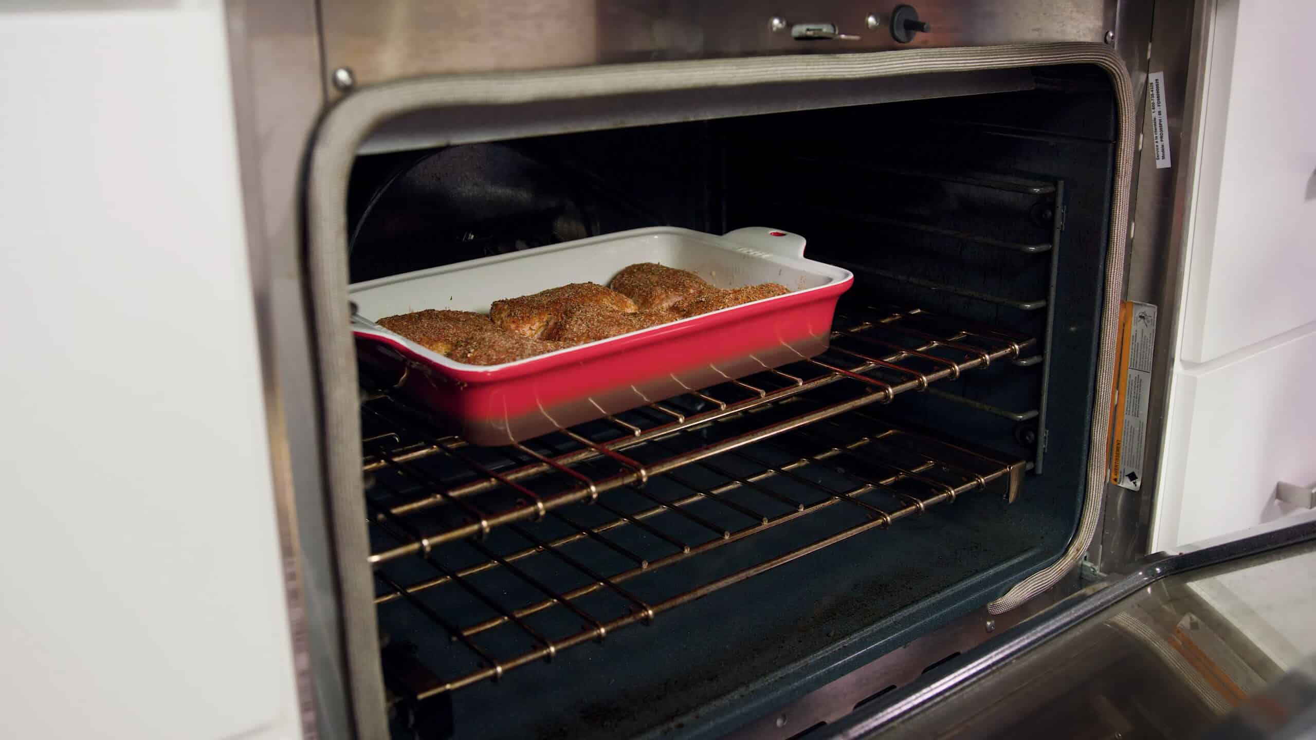 Angled view of open oven with an enamel coated cast iron casserole dish filled with chicken thighs placed on a metal rack in the middle of the oven.
