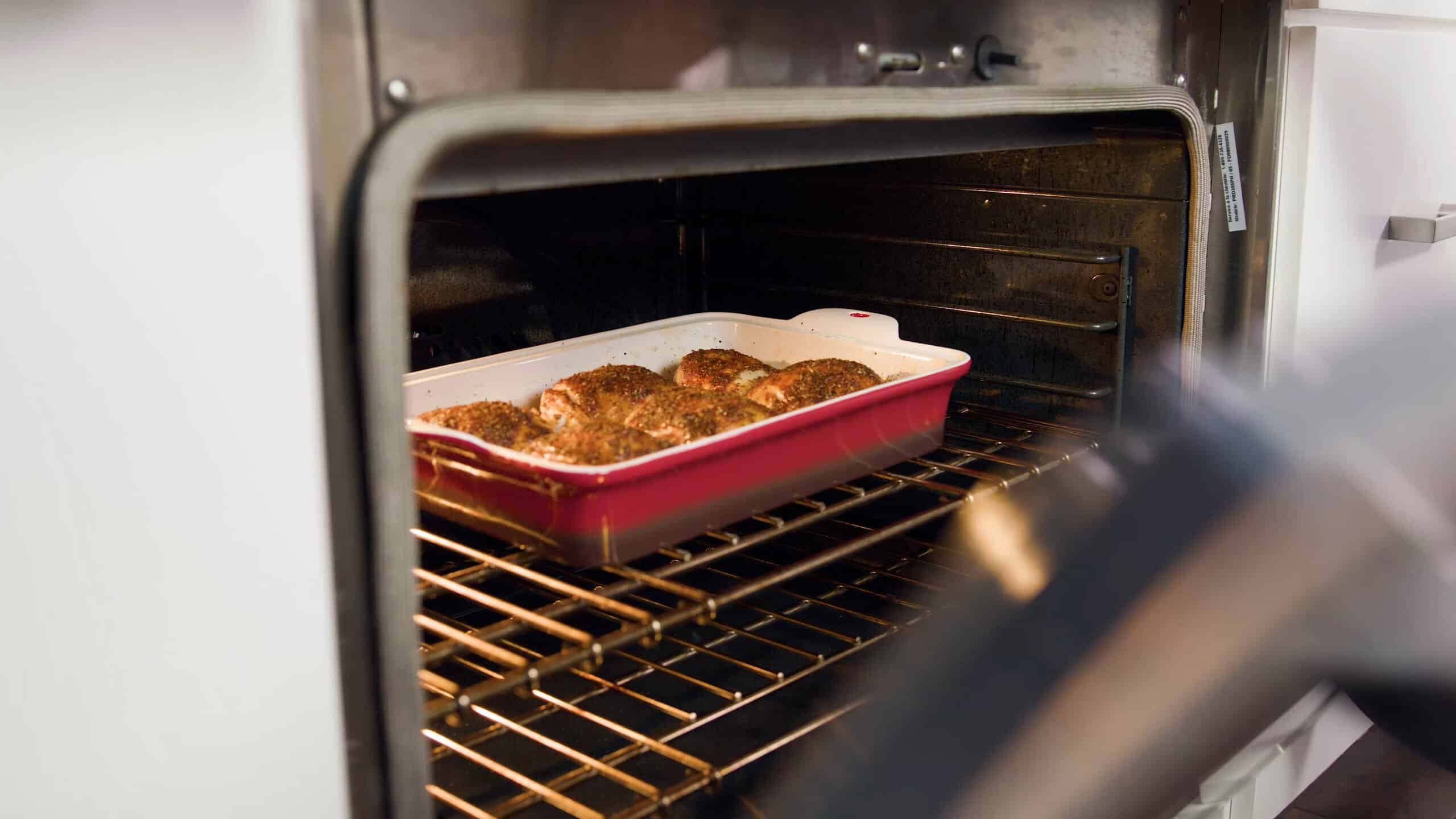 Angled view of open oven with an enamel coated cast iron casserole dish filled with chicken thighs placed on a metal rack in the middle of the oven for a second time.