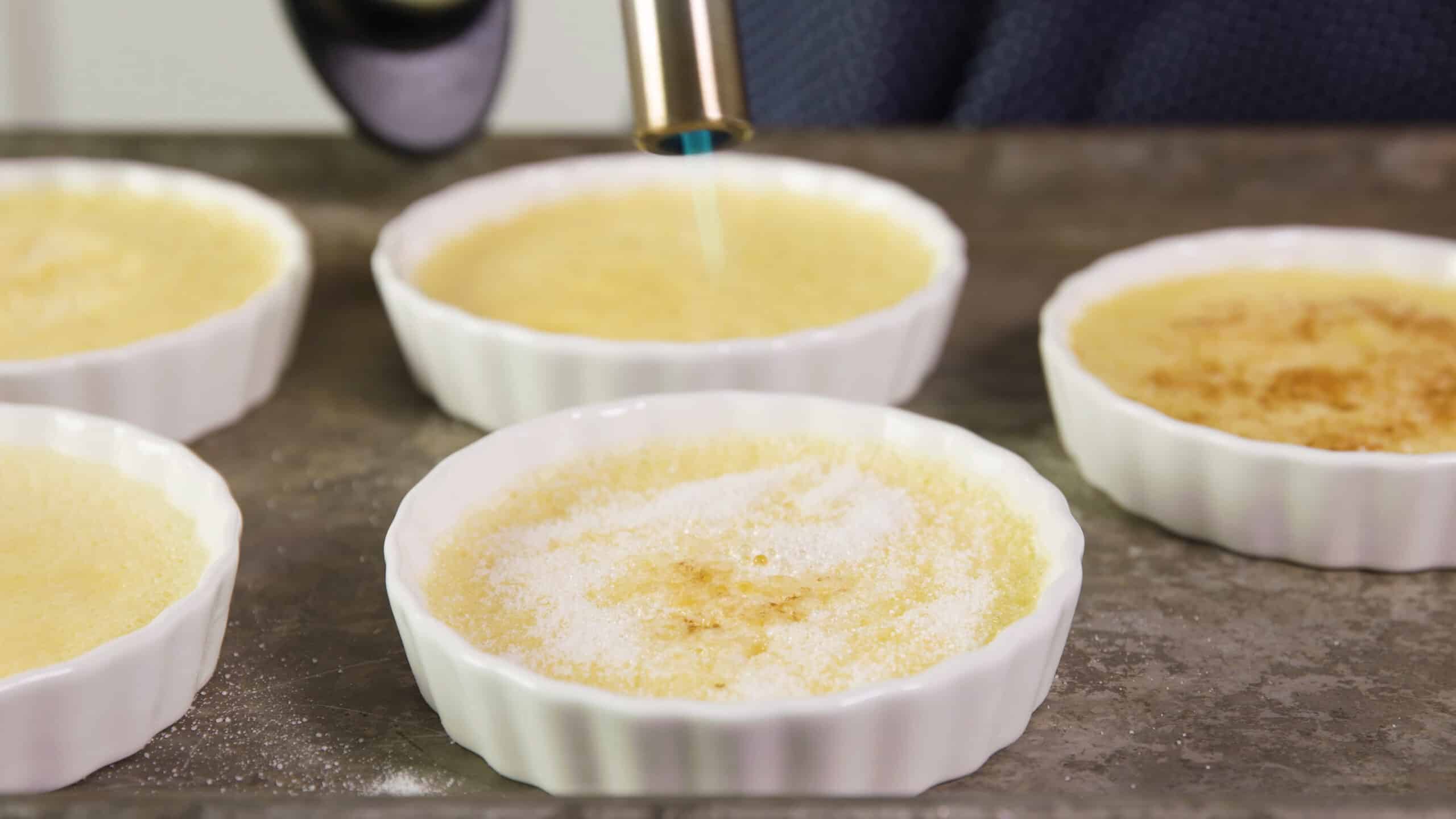 Angled view of metal baking sheet with white glass ramekins filled with chilled egg mixture and being topped with sugar the top being cooked/toasted with a creme brûlée torch.