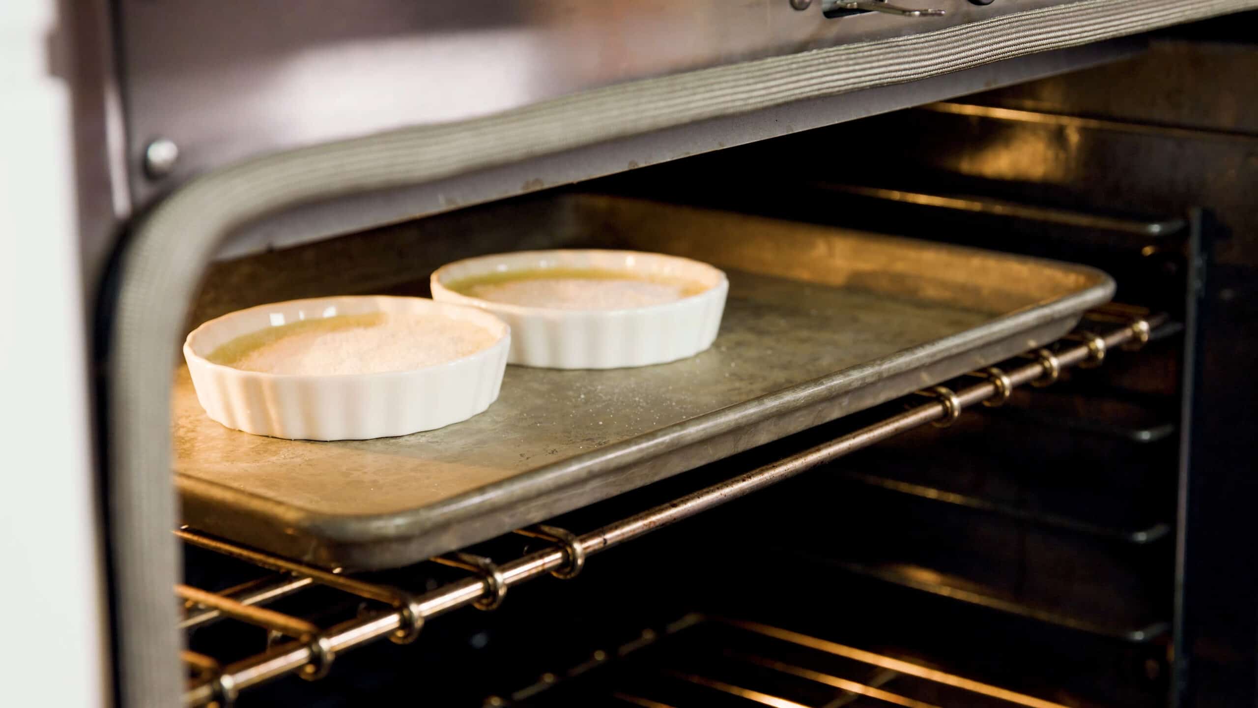 Angled view of open oven with a metal baking dish with two white glass ramekins filled with chilled egg mixture covered with white granulated sugar on a metal rack raised to the top level just under the broiling element of the oven.