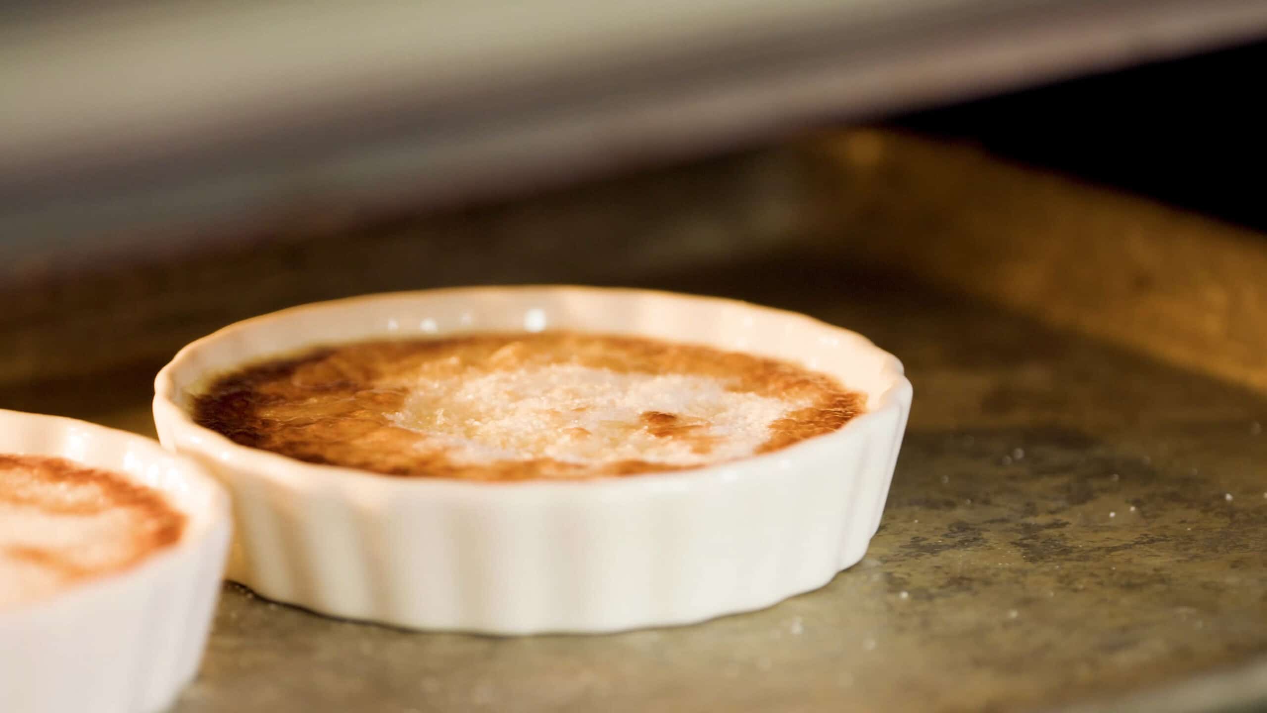 Close-up view inside an open oven showing a white glass ramekin filled with creme brûlée setting on a metal baking sheet white broiled to create the caramelized encrusted top of the dessert.