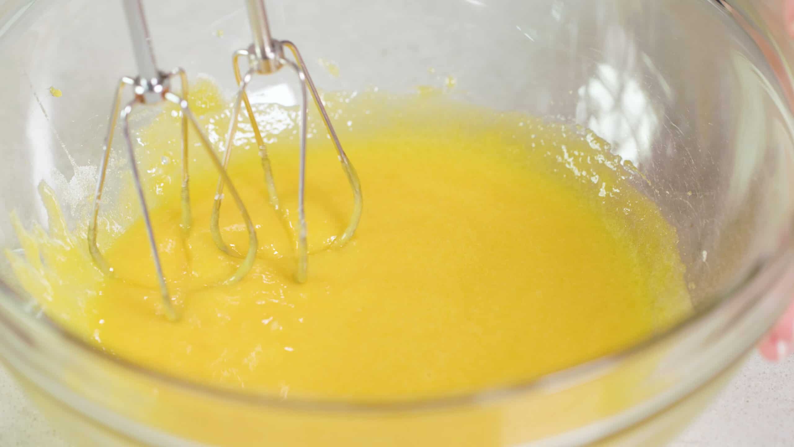 Close-up view of beaten egg yolks in a large clear glass mixing bowl with the metal wire beaters from a hand mixing.