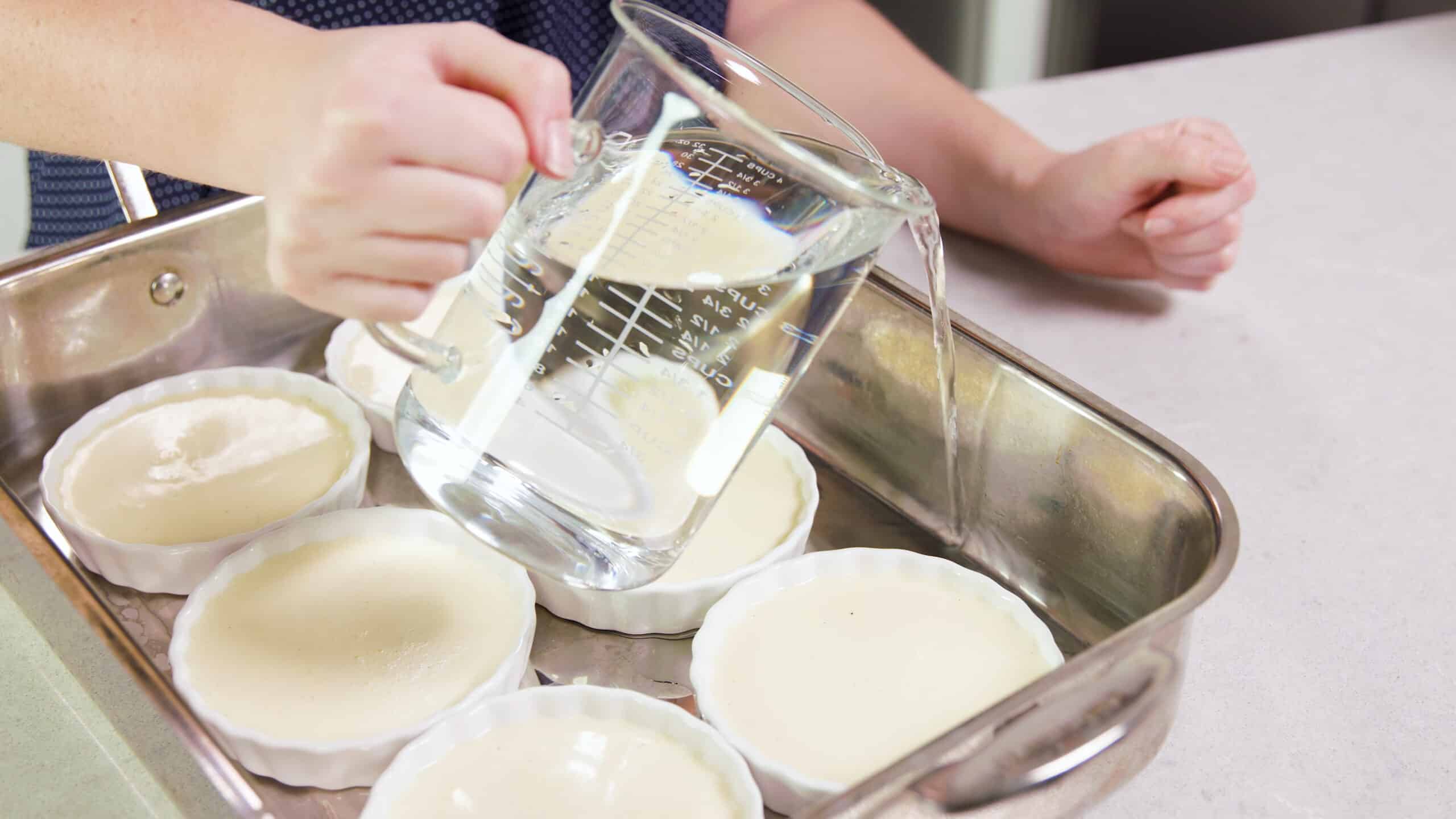 Angled view of a large metal hotel pan filled with six white glass ramekins filled with strained egg mixture and a large clear glass measuring cup pouring water into the spaces between each ramekin.