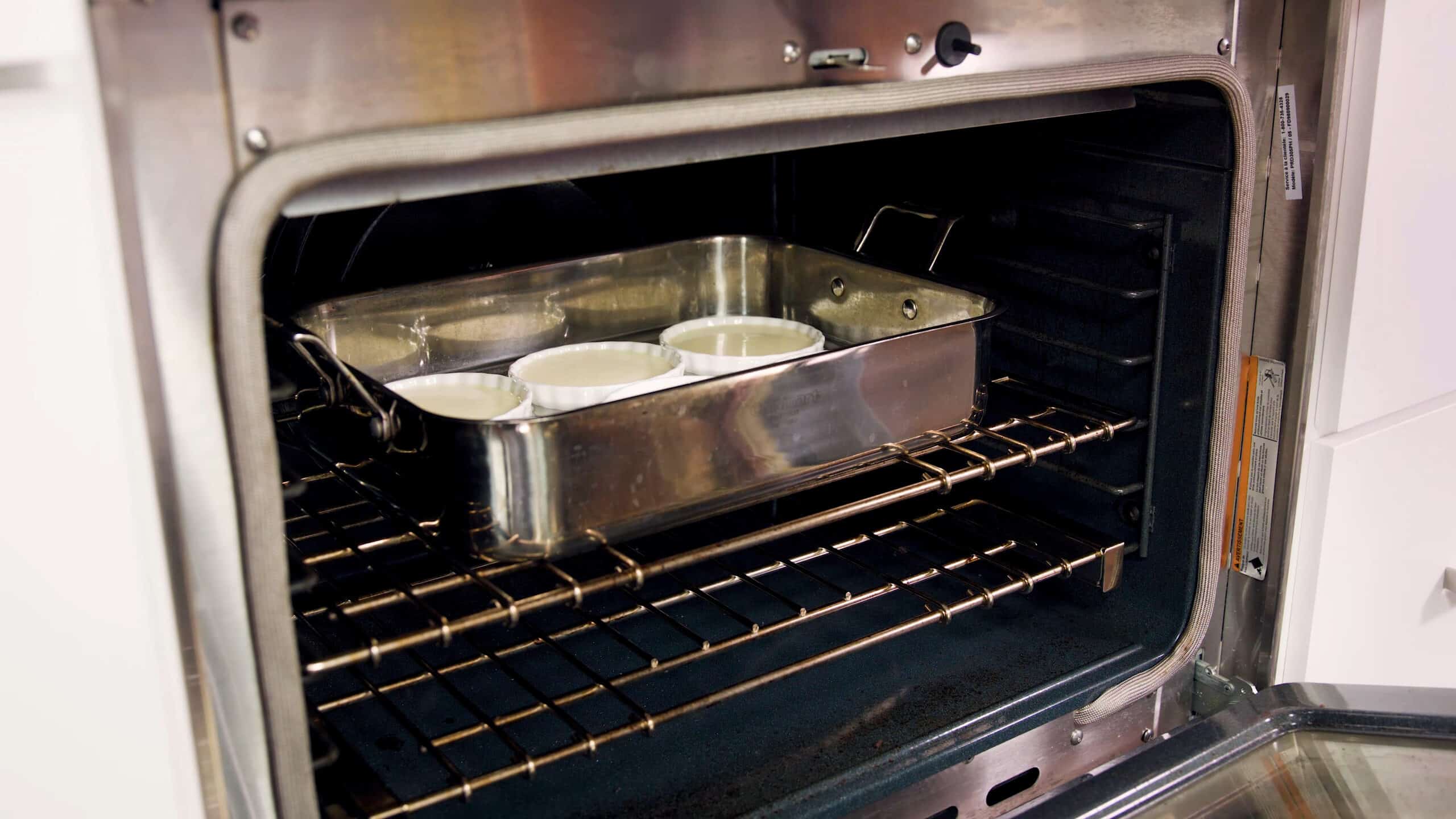Angled view of open oven with a large metal hotel pan filled with six white glass ramekins filled with strained egg mixture floating in a water bath all placed on the metal rack in the middle of the oven.