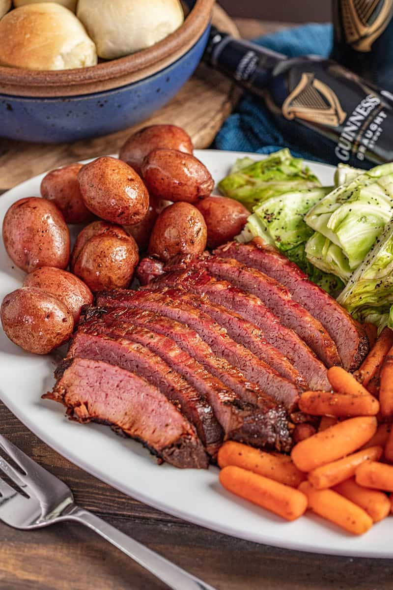 Sliced corned beef and cabbage with carrots and potatoes on platter with Guinness and fresh baked rolls in the background