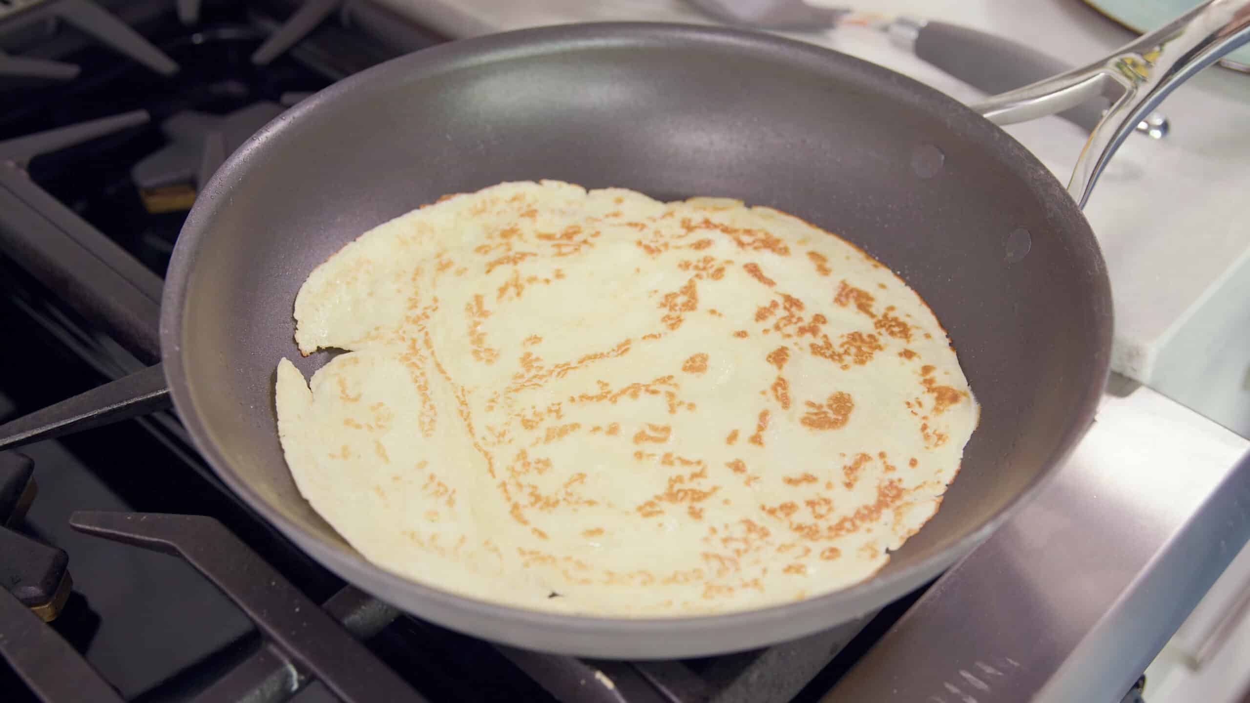 Angled view of metal non-stick pan with a flipped crepe showing the cooked side with golden brown areas cooked to perfection all over a stovetop burner.
