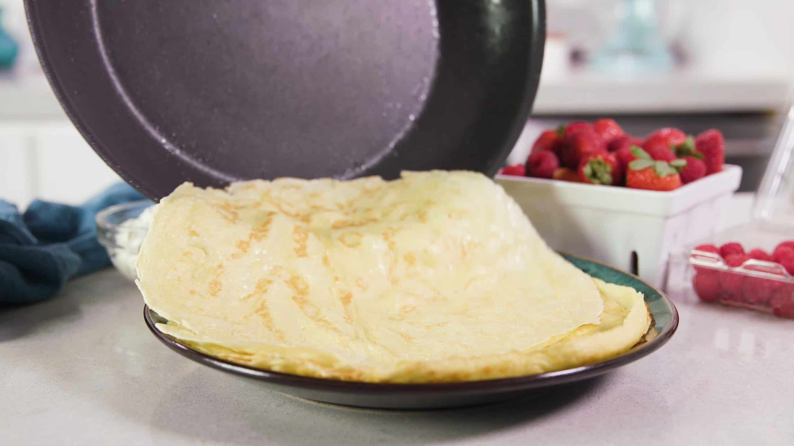 Angled view of a ceramic green and brown plate holding warm crepes with a freshly cooked crepe sliding on from a metal non-stick pan.