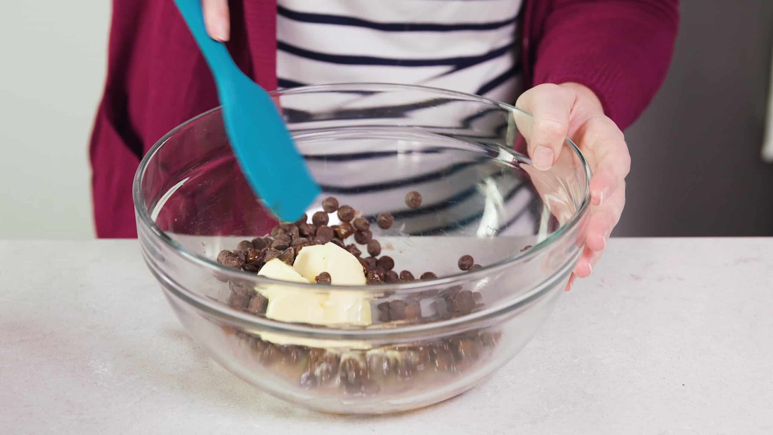 Angled view of large clear glass mixing bowl with melting chocolate chips and melting butter being stirred together with a plastic spatula.
