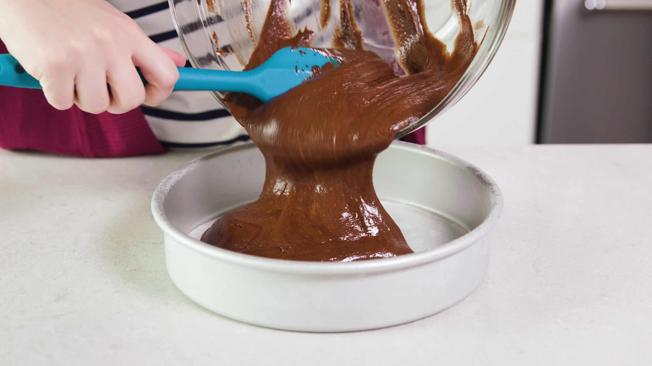 Angled view of metal round cake pan with cake batter being poured from a large clear glass mixing bowl scooped out with a plastic spatula.