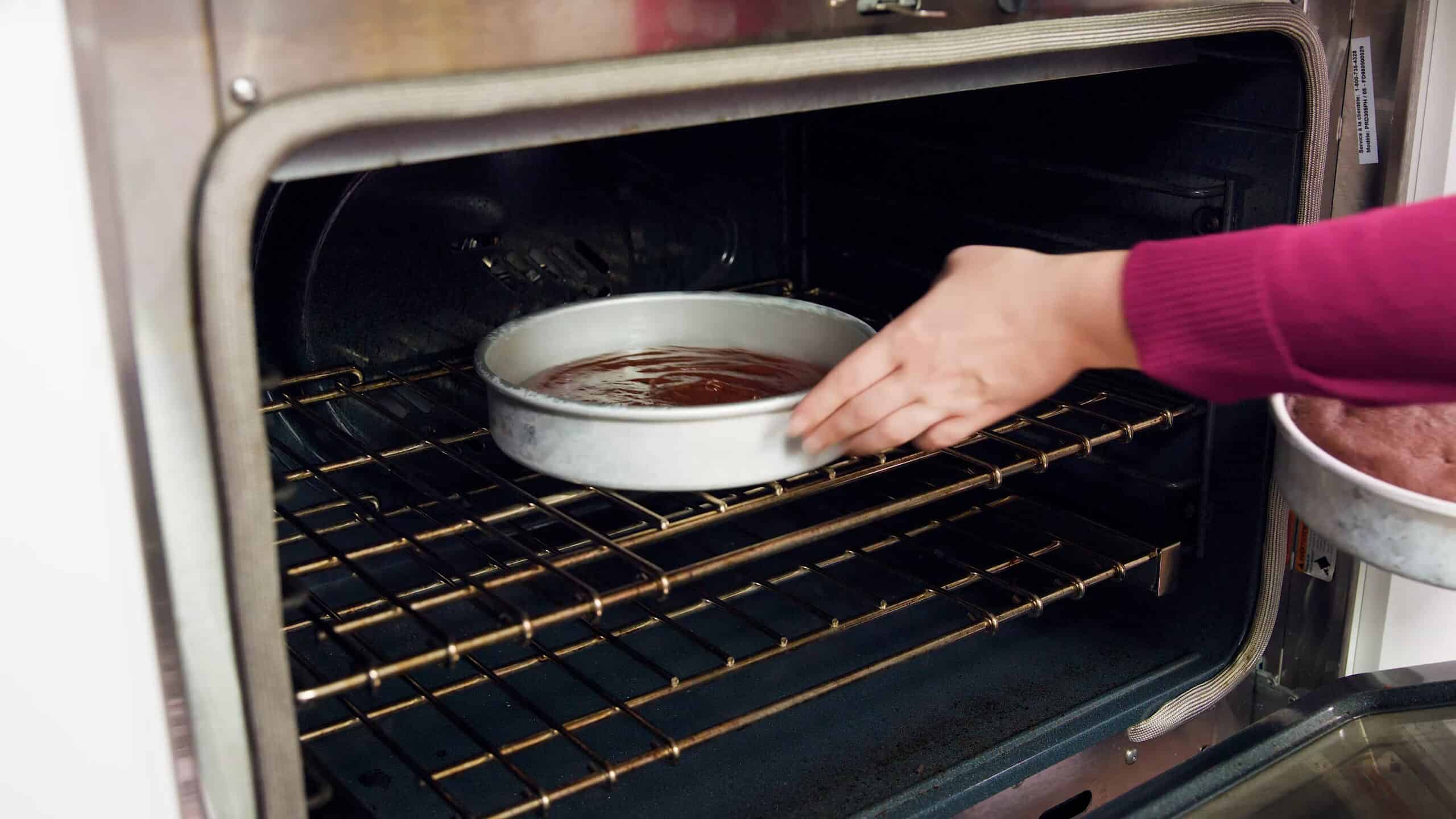 Angled view of an open oven with a metal round cake pan filled with chocolate cake batter placed on a metal rack in the middle of the oven.