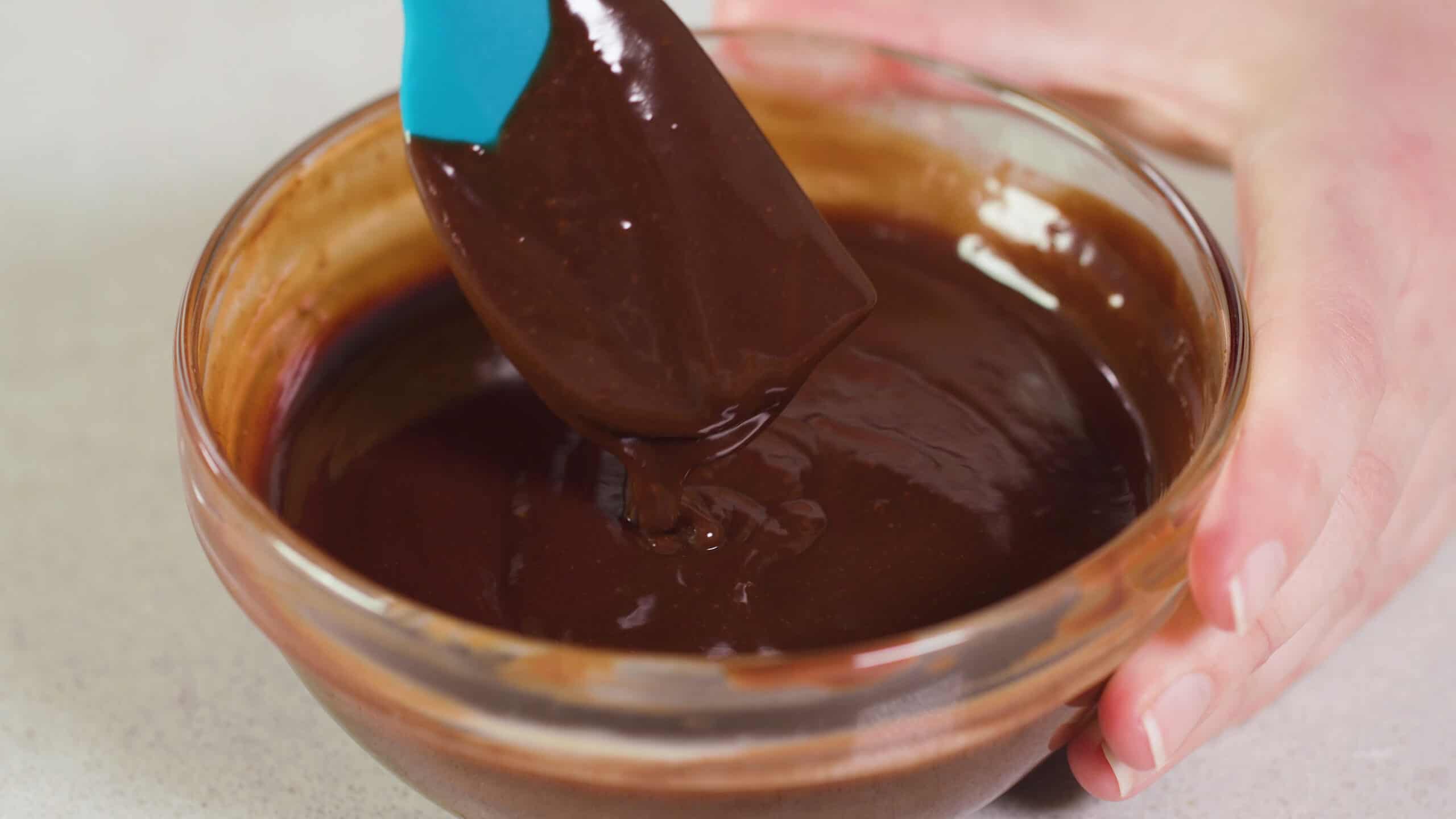 Close-up view of medium sized clear glass mixing bowl filled with rich chocolate frosting also dripping from a plastic spatula.
