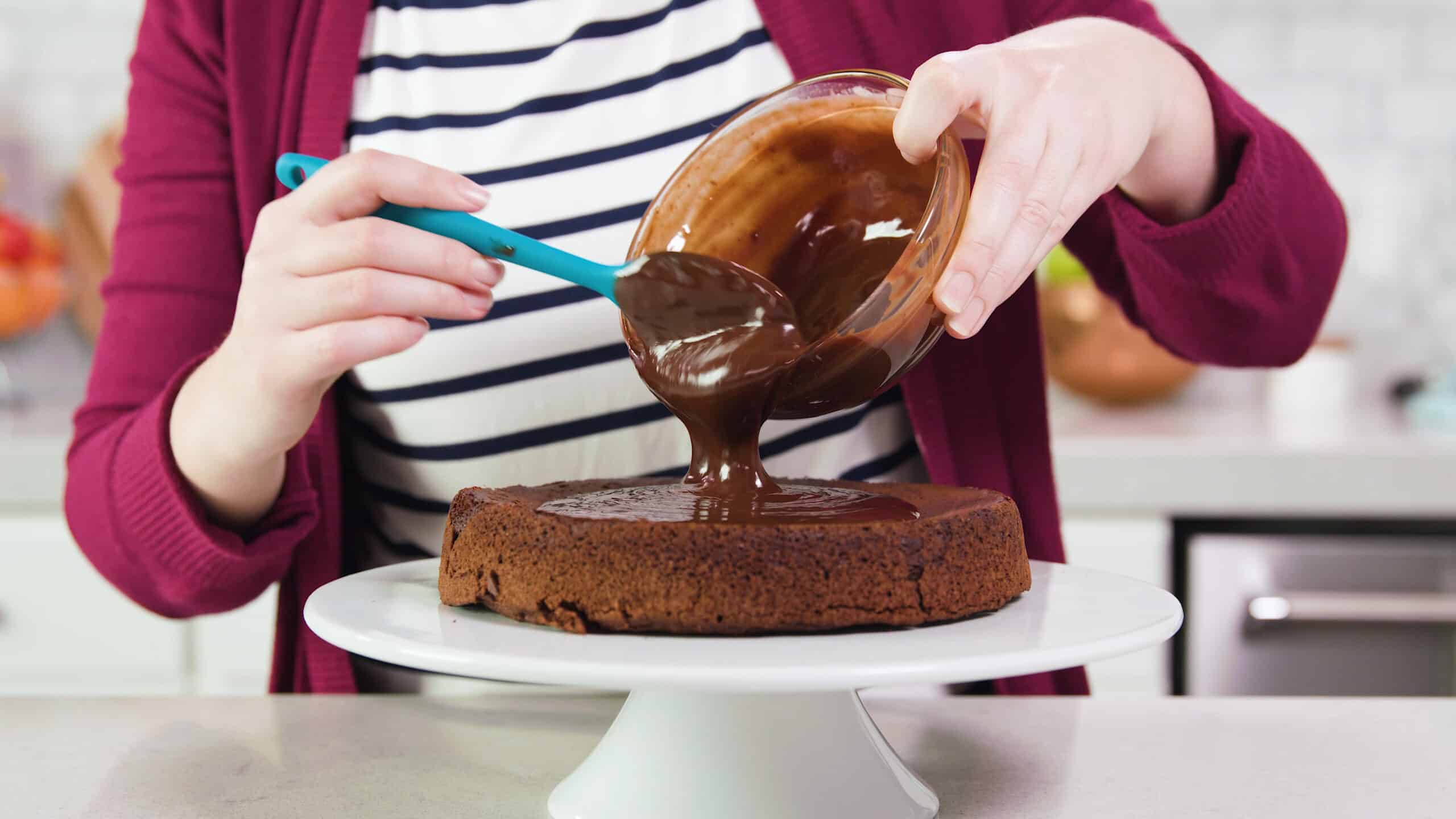 Side view of a white glass cake platter with baked flourless chocolate round cake on top with glossy chocolate frosting being pour from a clear glass mixing bowl scooped out with a plastic spatula.