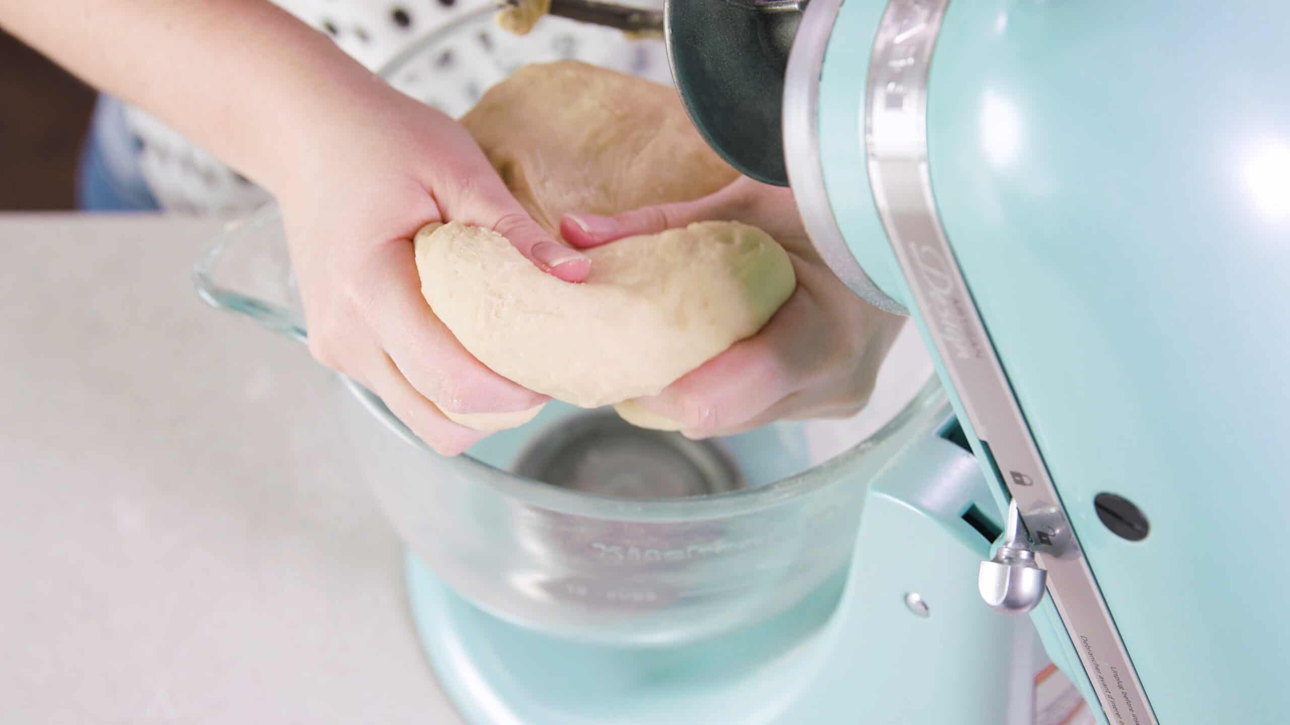 Overhead view of countertop kitchen mixer with clear glass mixing bowl and the kneaded dough held in hand to show texture.