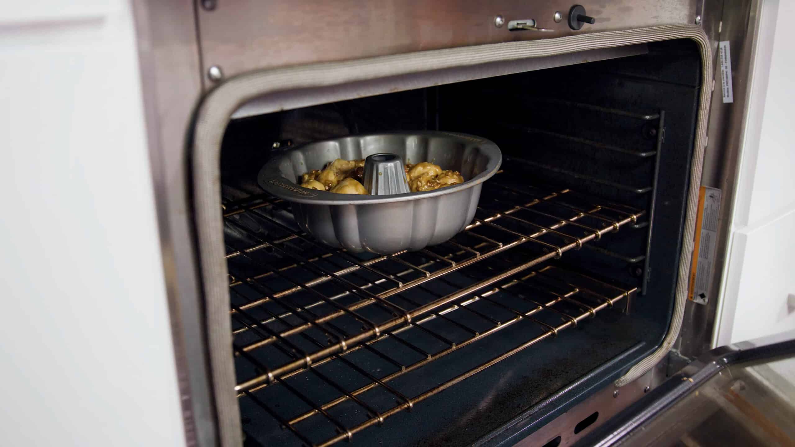 Angled view of an open oven with a metal bundt shaped cake pan filled with risen dough balls covered in pecan caramel sauce sitting on a metal rack in the middle of the oven.