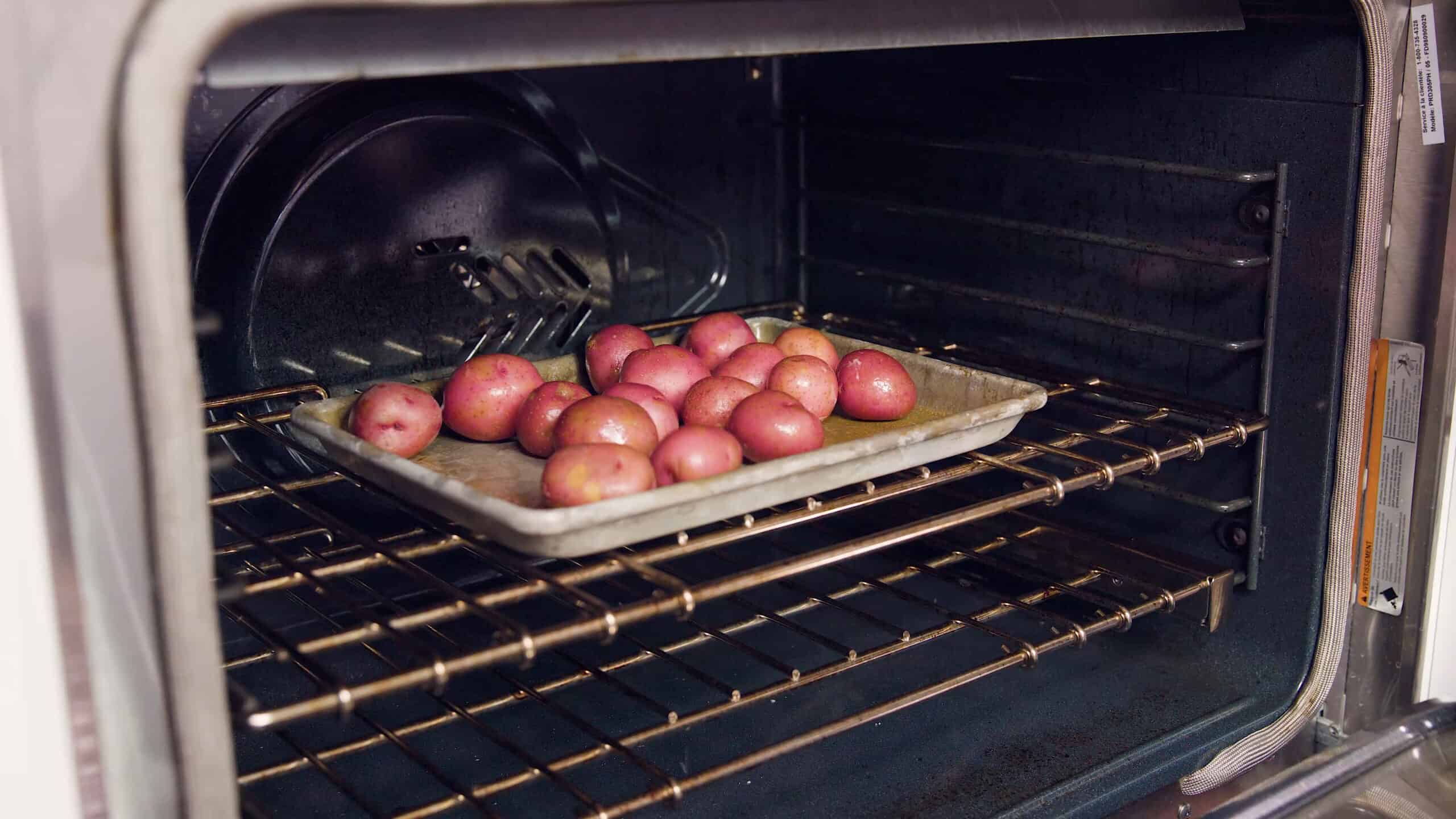 Angled view of open oven with a metal baking sheet with about one dozen red potatoes placed on a metal rack in the middle of the oven.
