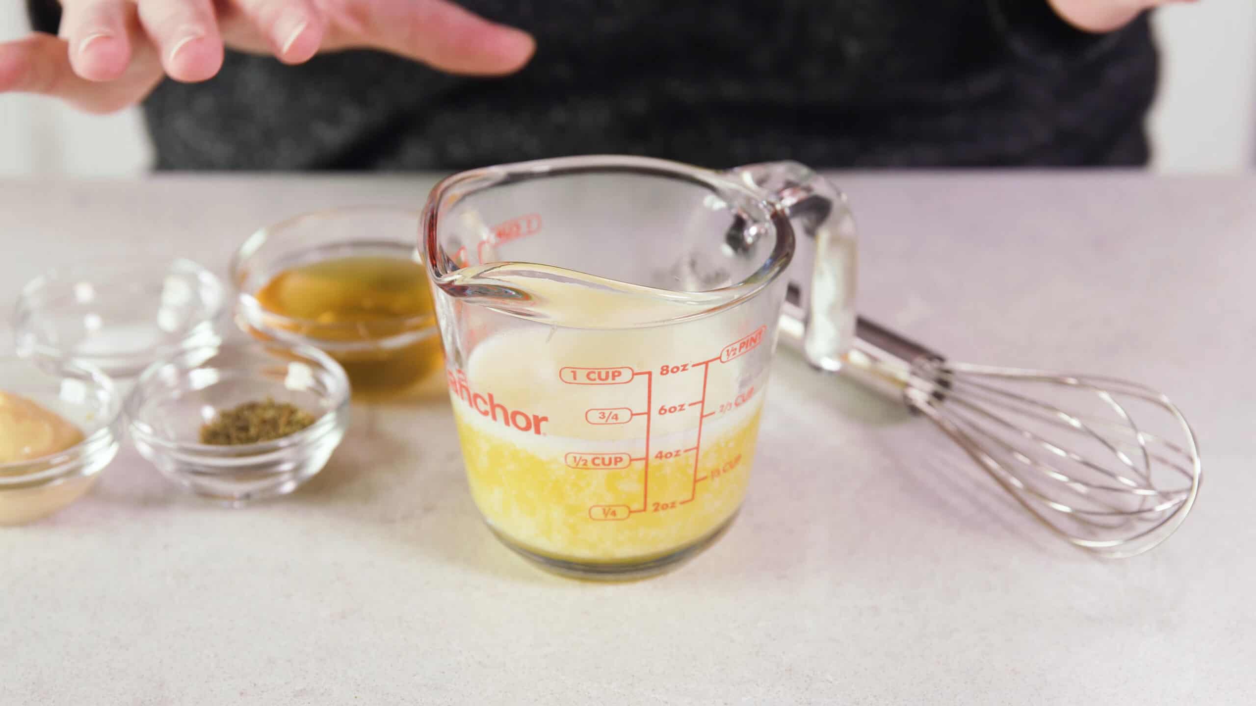 Angled view of clear glass measuring cup filled with melted butter and other wet and dry ingredients in smaller clear glass mixing bowls to the left and a wire whisk to the right.