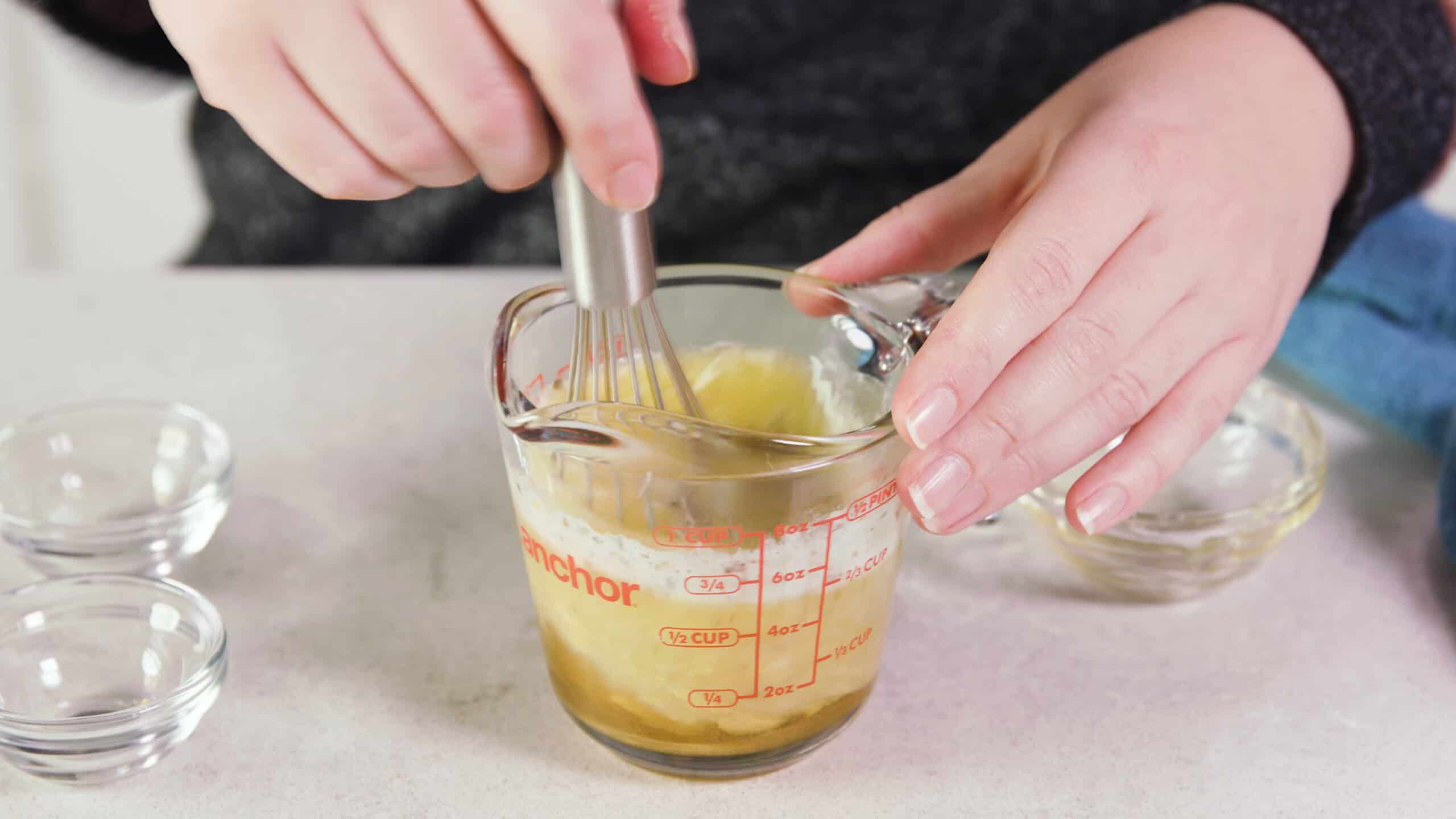 Angled view of clear glass measuring cup filled with melted butter and other ingredients for flavoring and a wire whisk mixing the ingredients together.