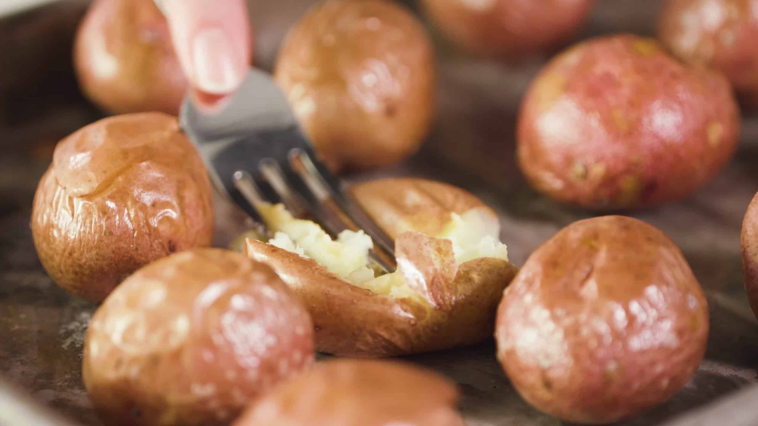 Close-up view of a metal fork smashing a single red potato amongst the remains red potatoes on a metal baking sheet.