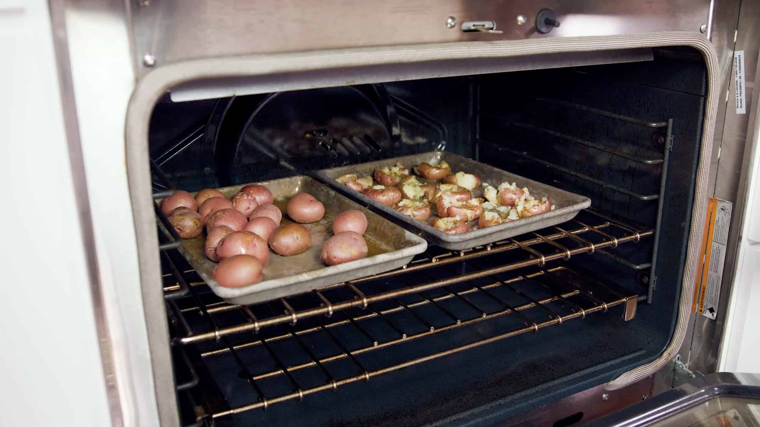 Angled view of open oven with two metal baking sheets, one of the left with whole red potatoes, and one on the right with smashed red potatoes with melted butter mixture poured on top all set on a metal rack in the middle of the oven.