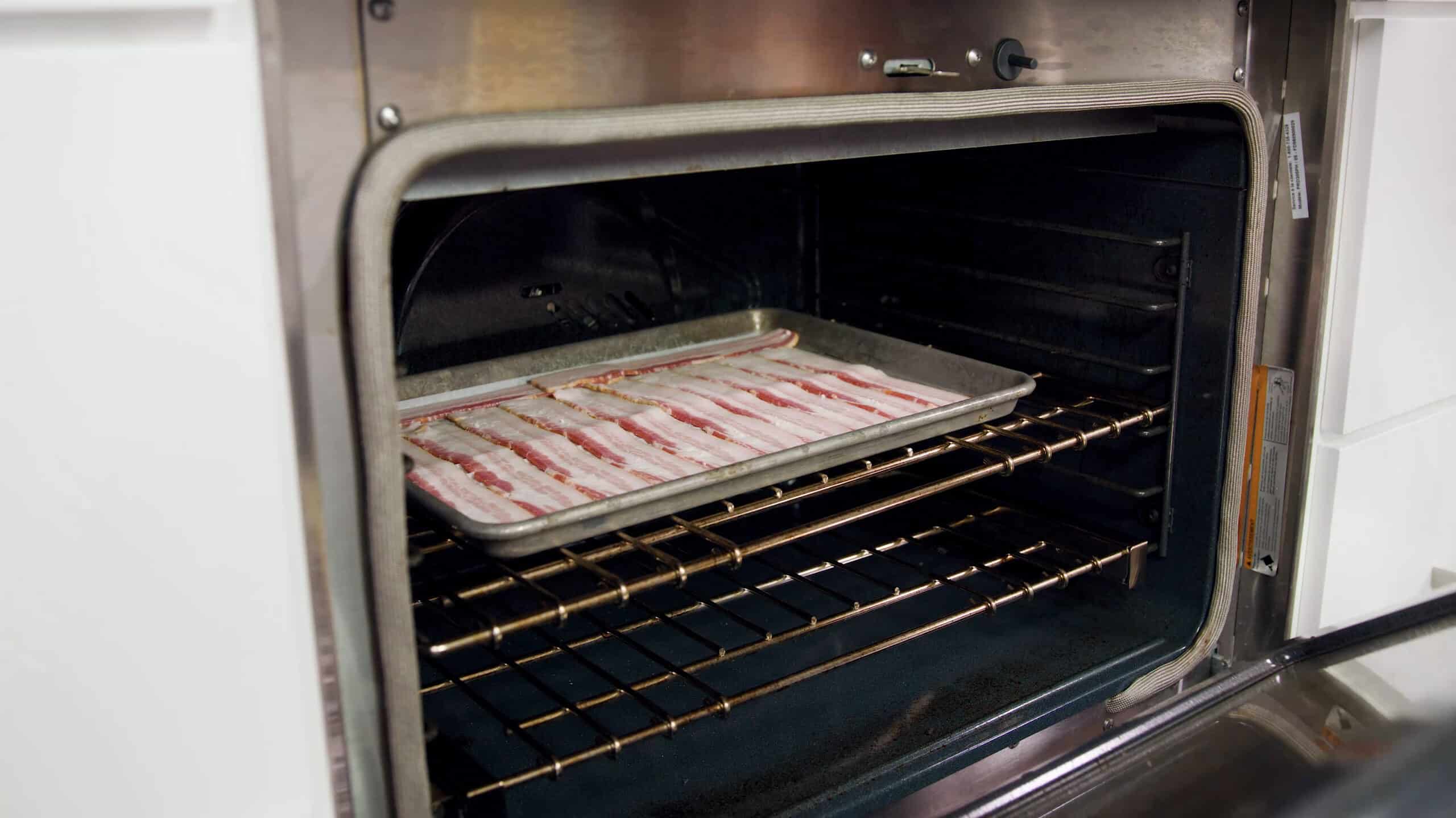 Angled view of an open oven with a metal baking sheet lined with parchment paper and lined with bacon placed on a. metal rack in the middle of the oven.