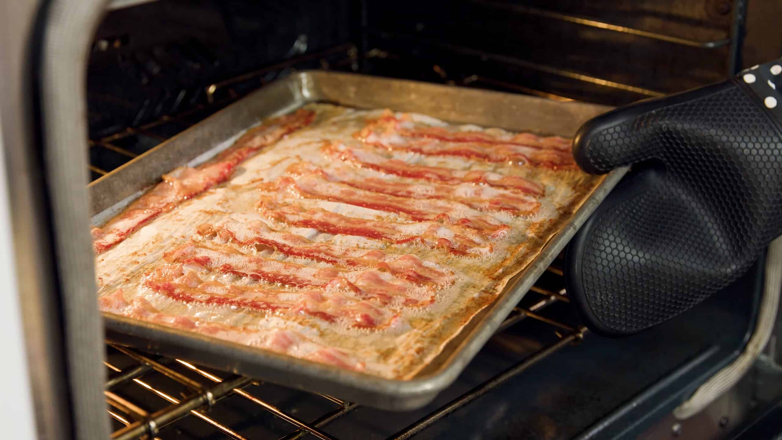 Angled view of an open oven with a metal baking sheet lined with parchment paper and cooking golden brown strips of bacon being pulled off of the metal rack in the middle of the oven.