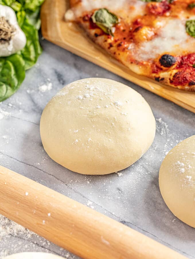 round pizza dough ball on a marble counter with a rolling pin and a cooked pizza in the background