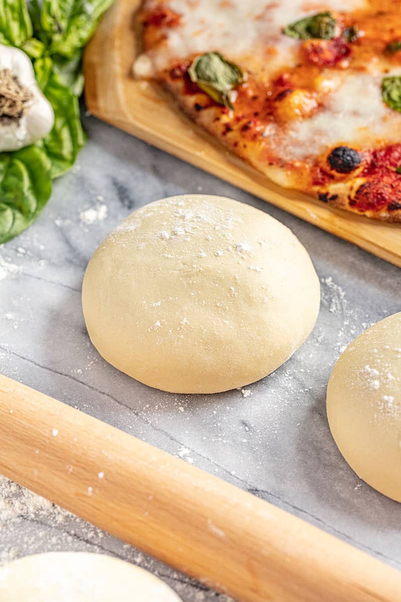 Pizza dough on a marble counter with a rolling pin and cooked pizza in the background