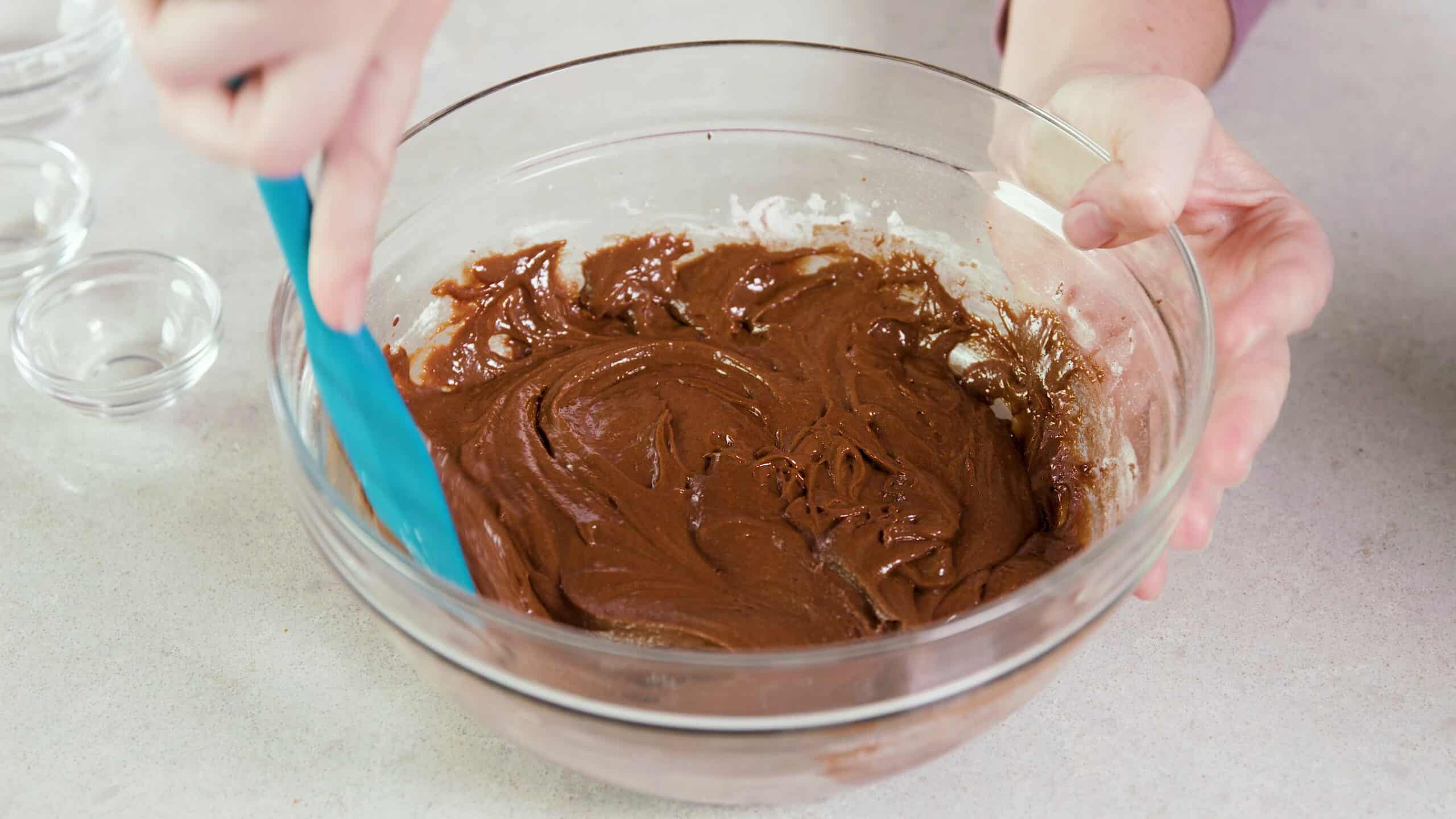 Angled view of large clear glass mixing bowl filled with brownie mix being stirred by a plastic spatula.