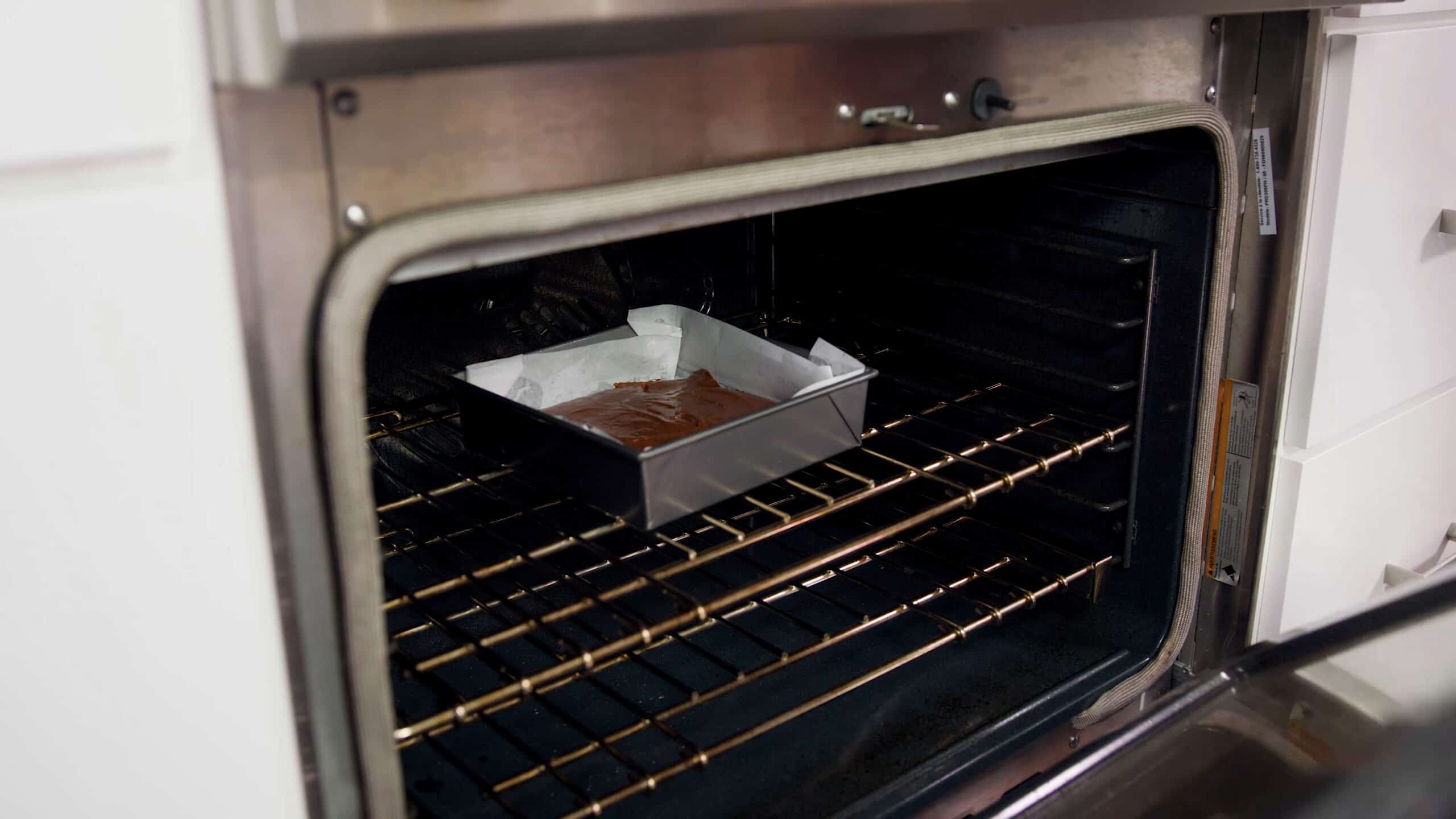 Angled view of open oven with a square metal brownie pan lined with parchment paper and filled with brownie mix placed on a metal rack in the middle of the oven.