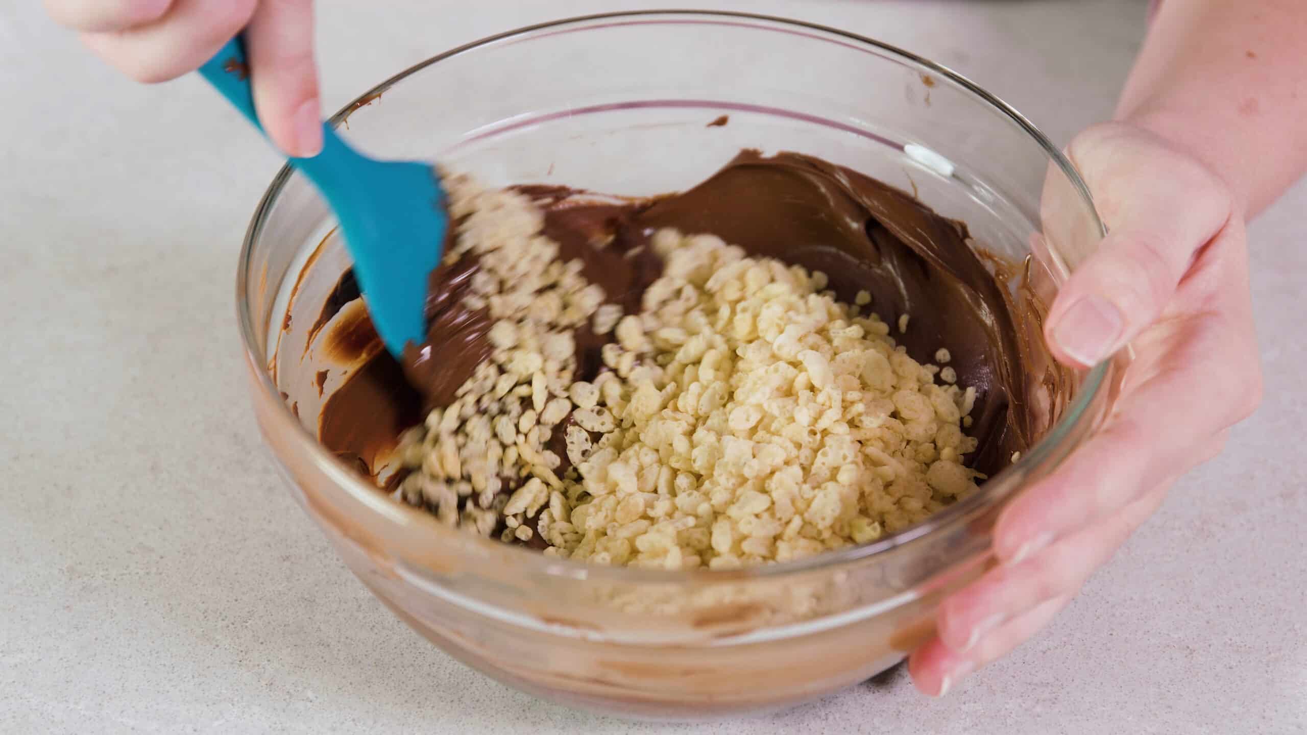 Angled view of large clear glass mixing bowl filled with melted chocolate and mixed with Rice Krispies Cereal.