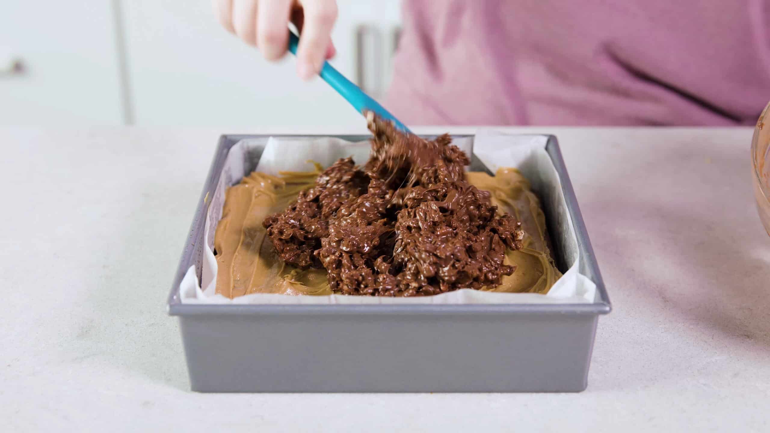Angled view of square metal brownie pan lined with parchment paper and filled with cooked brownies, a layer of peanut butter and topped with Rice Krispies Cereal and melted chocolate mixture, spread by a plastic spatula.