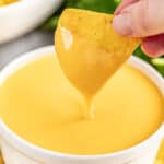 A hand dipping a tortilla chip into a small bowl of cheese sauce