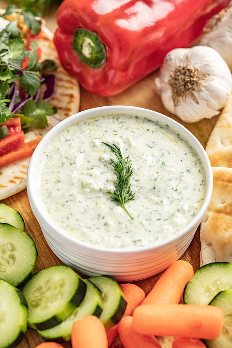 A bowl of tzatziki sauce garneshed with s sprig of dill, surrounded by fresh cucumber slices and baby carrots