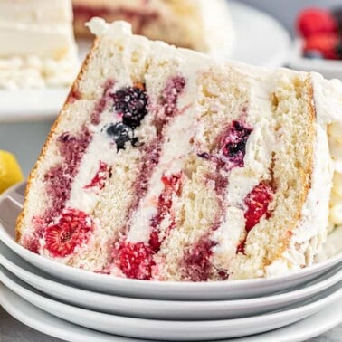 A slice of fresh berry Chantilly cake on a stack of dessert plates with fresh berries and lemon halves in the background