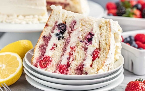A slice of fresh berry Chantilly cake on a stack of dessert plates with fresh berries and lemon halves in the background