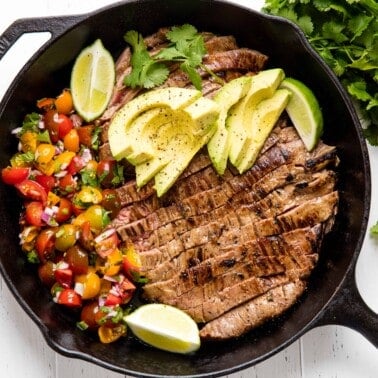 Bird's eye view of Mexican skillet steak in a skillet with sliced avocado, lime wedges, and sliced tomatoes.