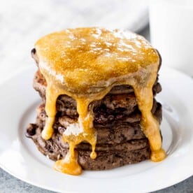 A stack of chocolate pancakes covered in a caramel syrup on a white plate.