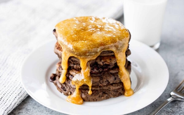 A stack of chocolate pancakes covered in a caramel syrup on a white plate.